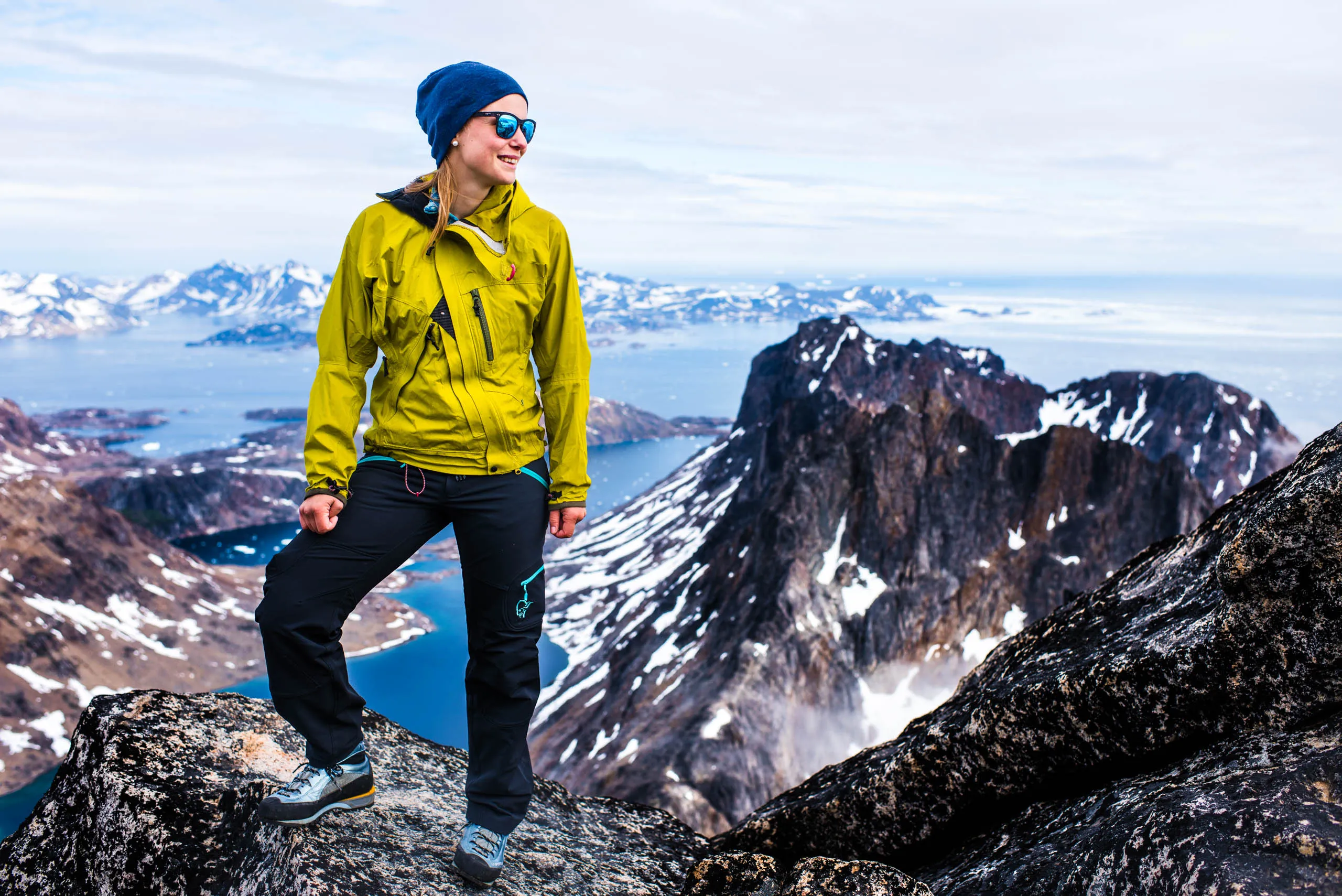 Smiling woman in yellow jacket and blue beanie resting on a summit with Greenland's fjords behind her