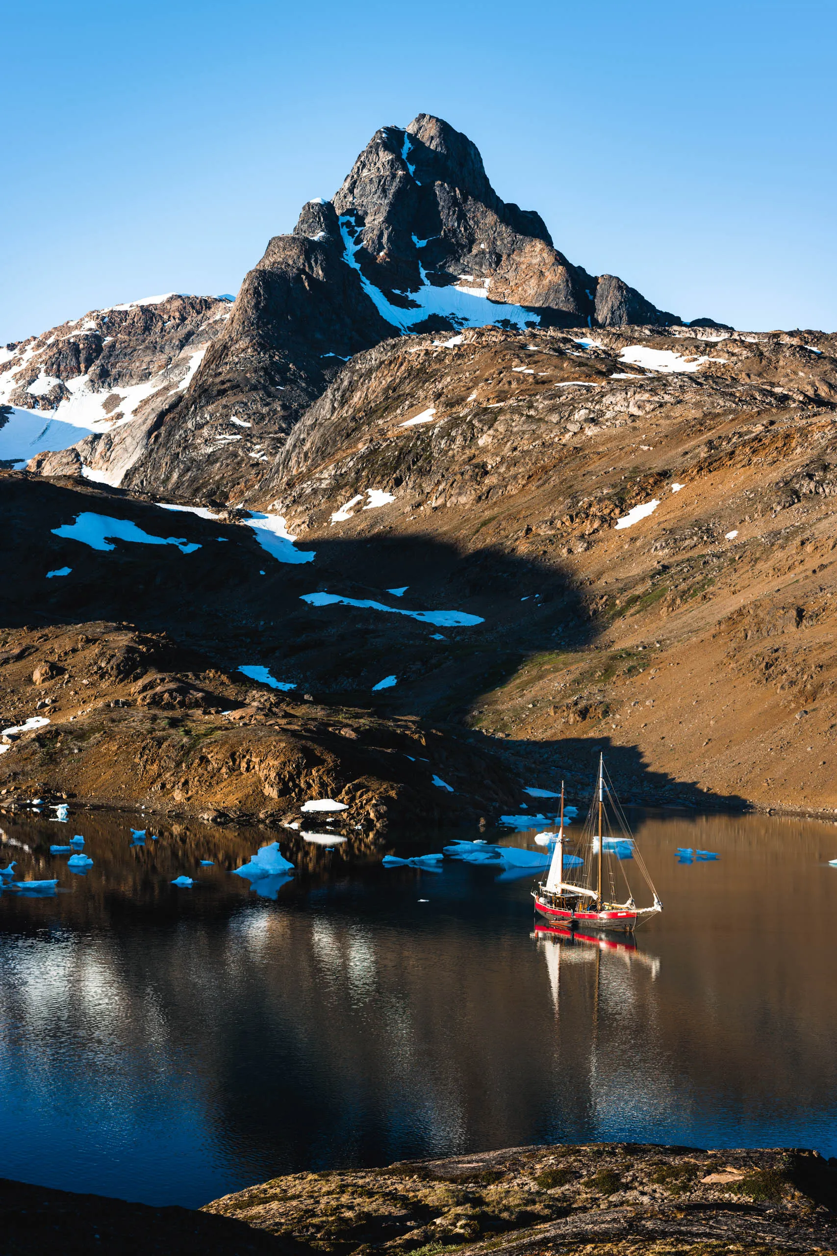 Red sailing vessel anchored in a calm fjord beneath a jagged Arctic peak with snow patches