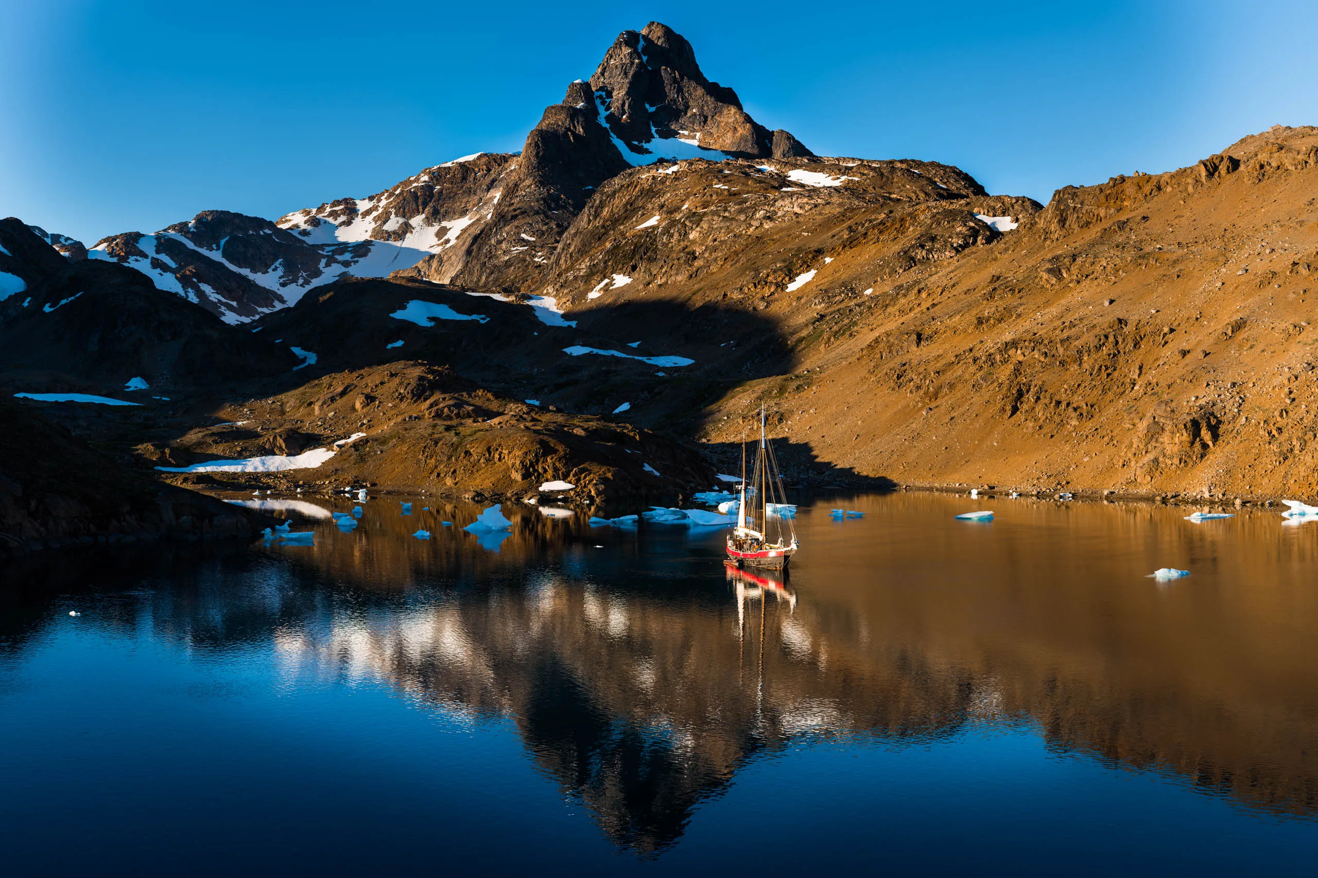 Red sailboat perfectly mirrored in a still fjord with small icebergs and a towering peak