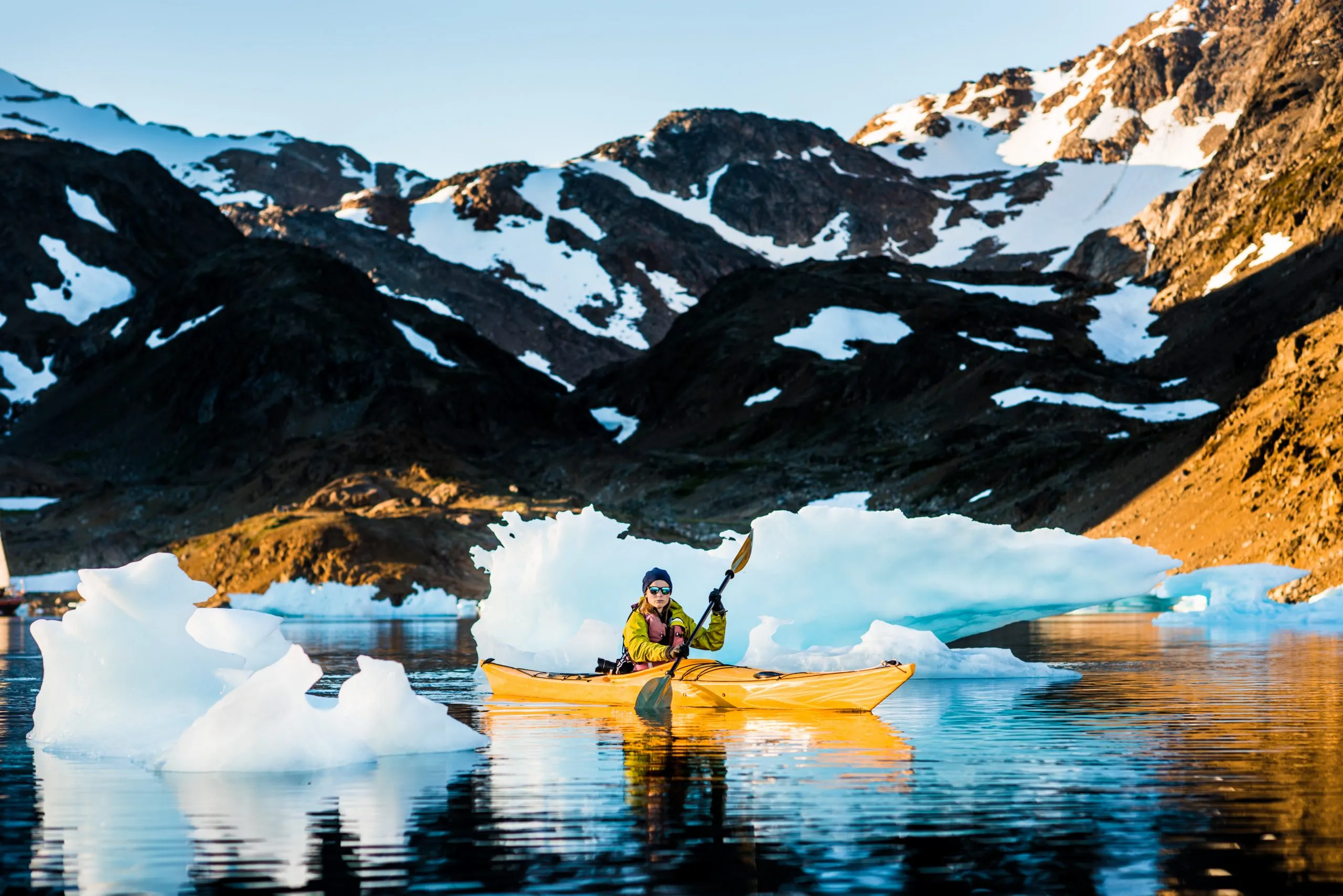 A person paddles a yellow sea kayak between drifting icebergs in front of snow-streaked mountains in East Greenland.