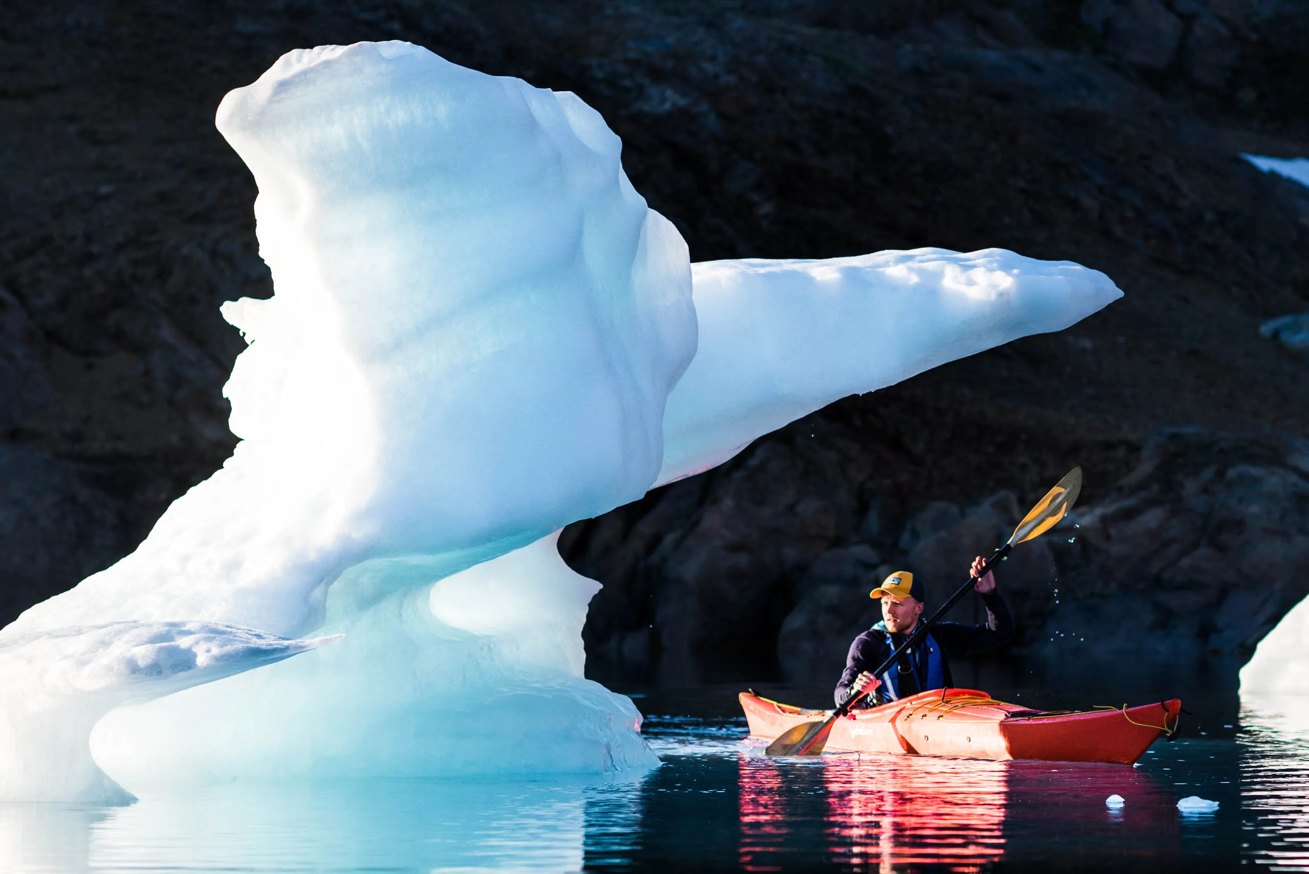 Kayaker in red boat dwarfed beside a luminous sculpted iceberg against dark rock