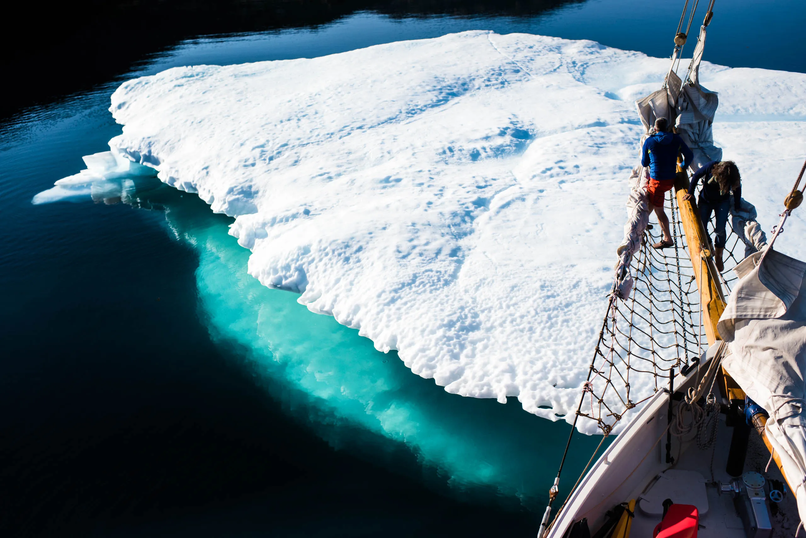 Crew members on the ship's bowsprit gazing at a massive flat iceberg in turquoise water