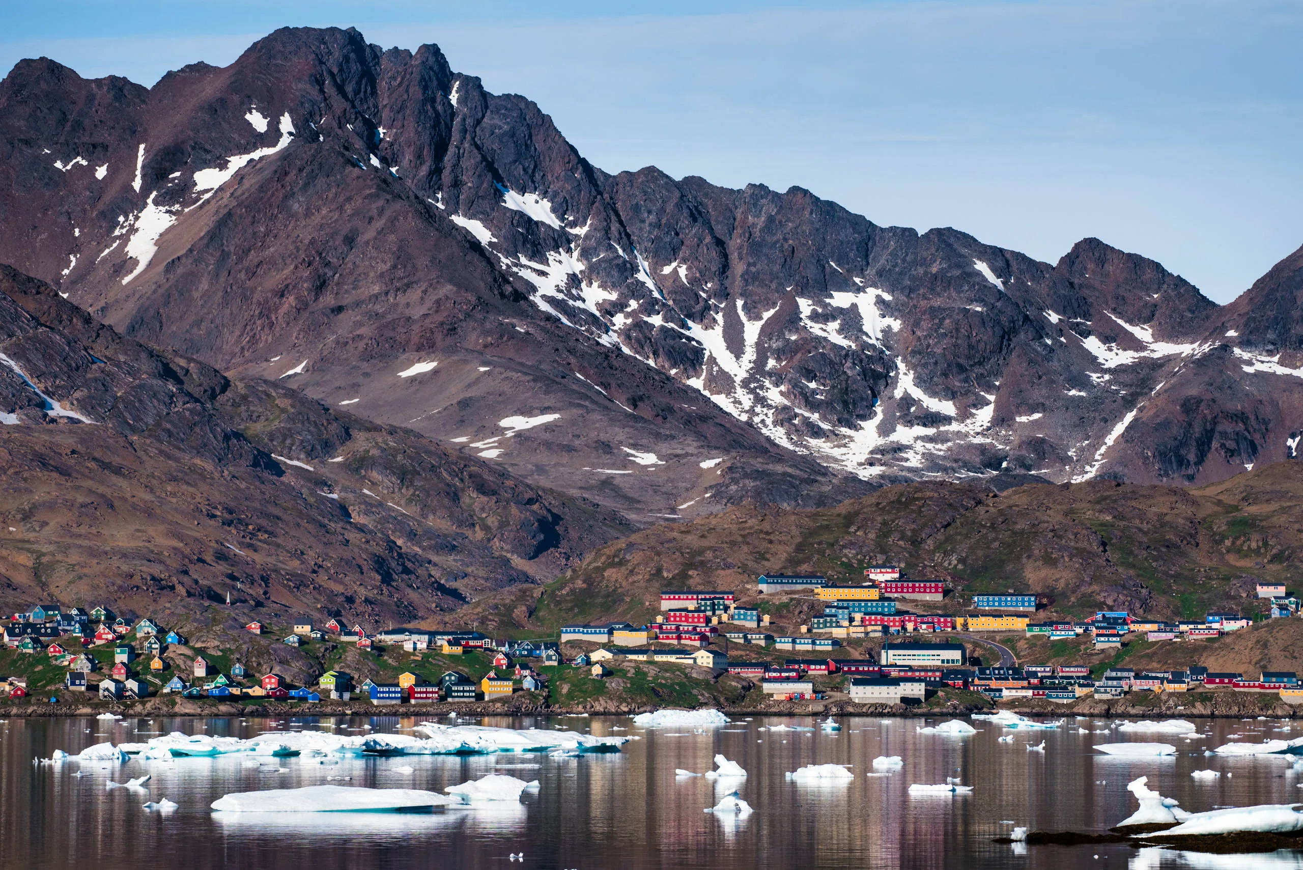 Colourful Greenlandic settlement reflected in icy fjord waters at the foot of vast snow-streaked mountains