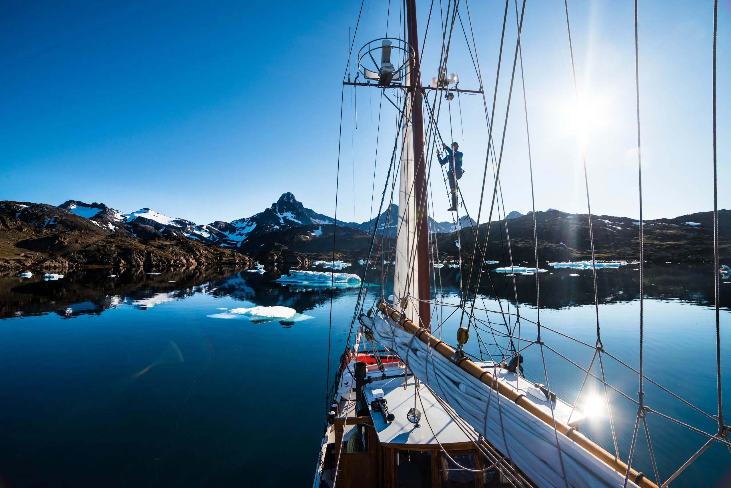 Crew member climbs the mast of a sailboat among icebergs in a calm Greenlandic fjord