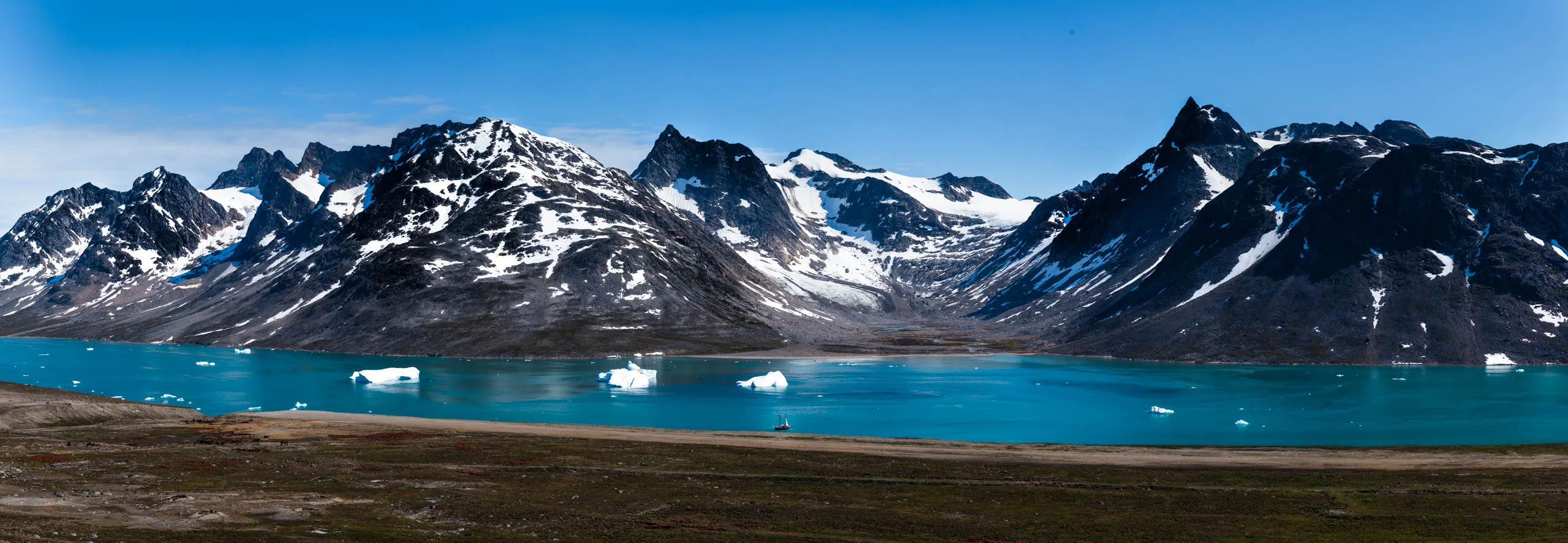Panoramic view of a turquoise glacial bay ringed by snow-patched mountains with a lone sailboat
