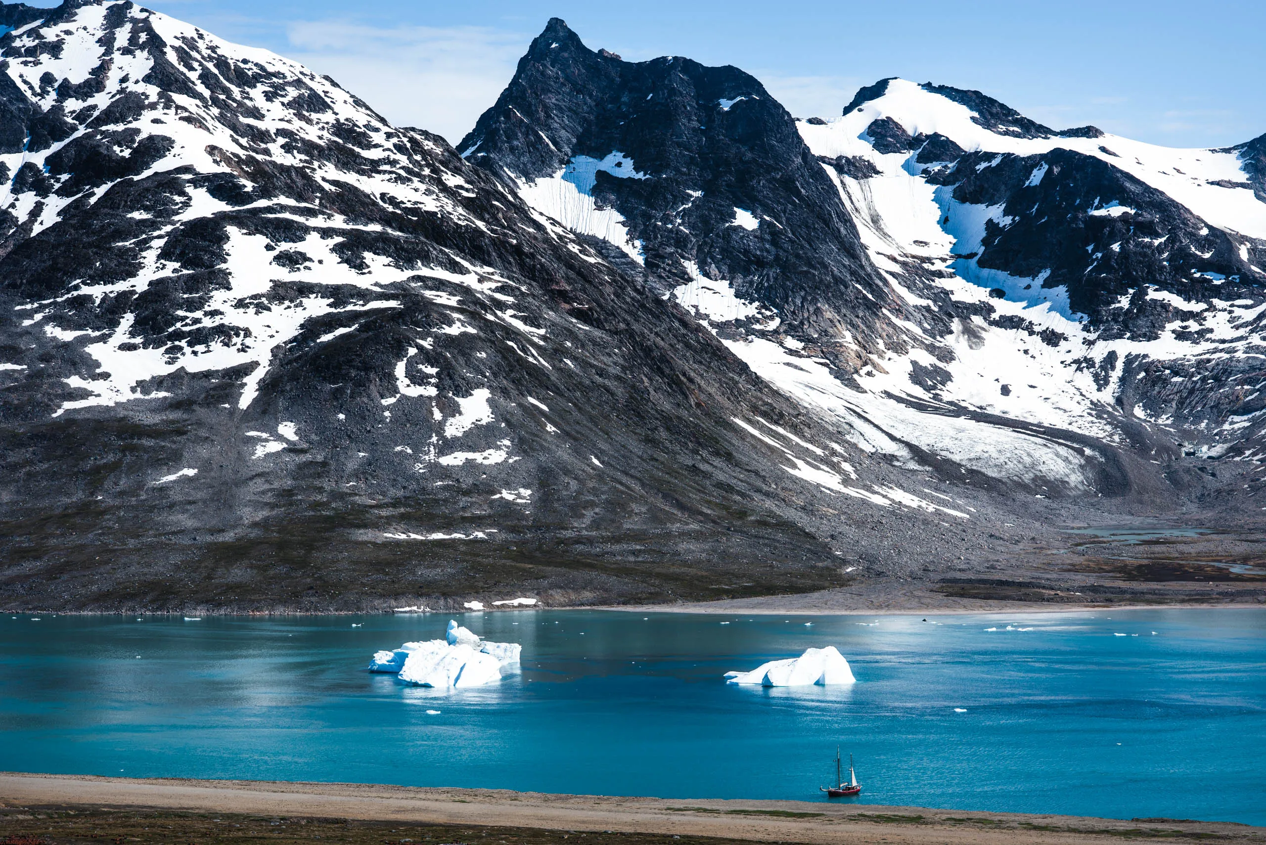 Small sailboat dwarfed by snow-streaked mountains and bright blue glacial water with floating icebergs