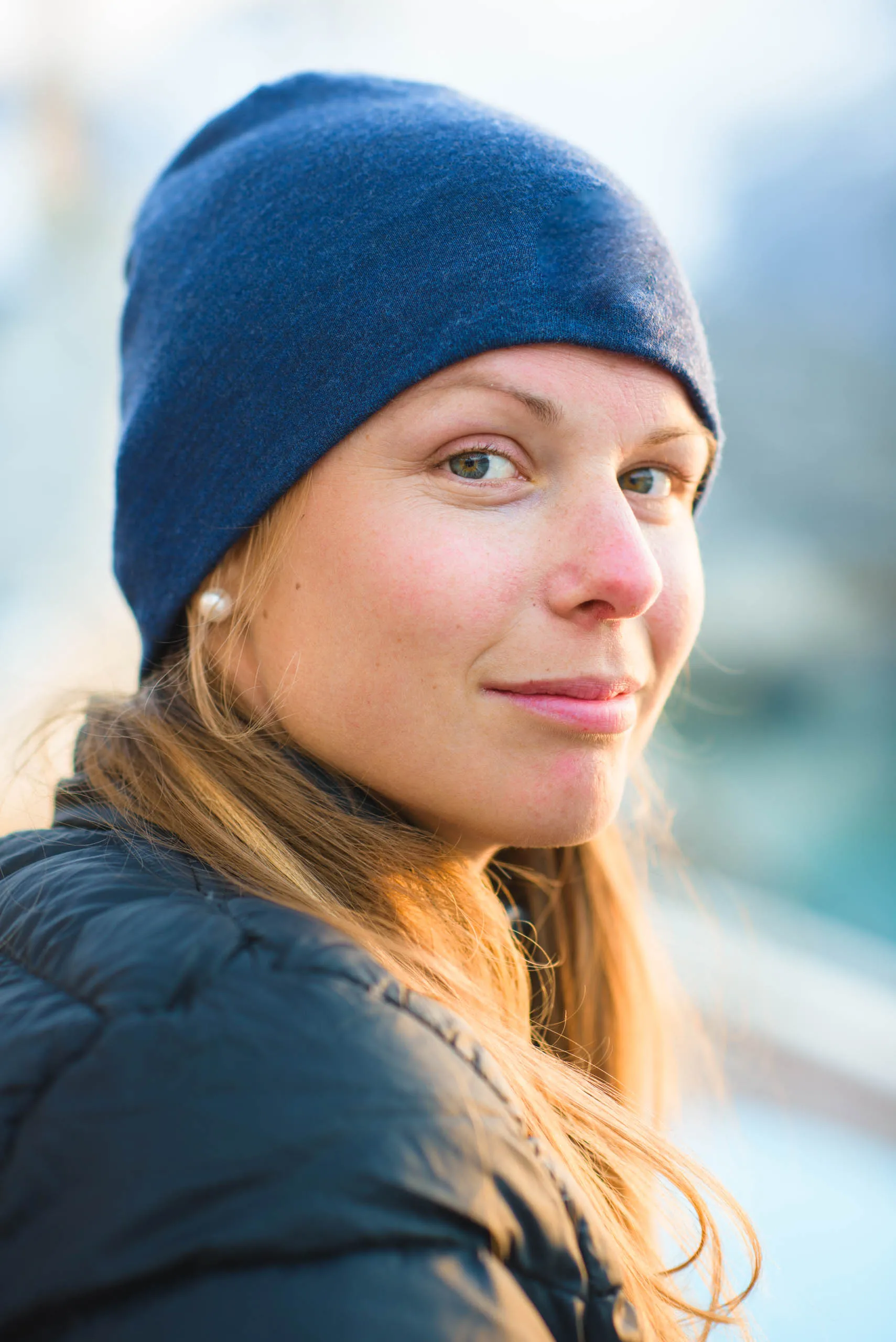 Close portrait of a young woman in a blue beanie, warm light on her face against a blurred Arctic backdrop