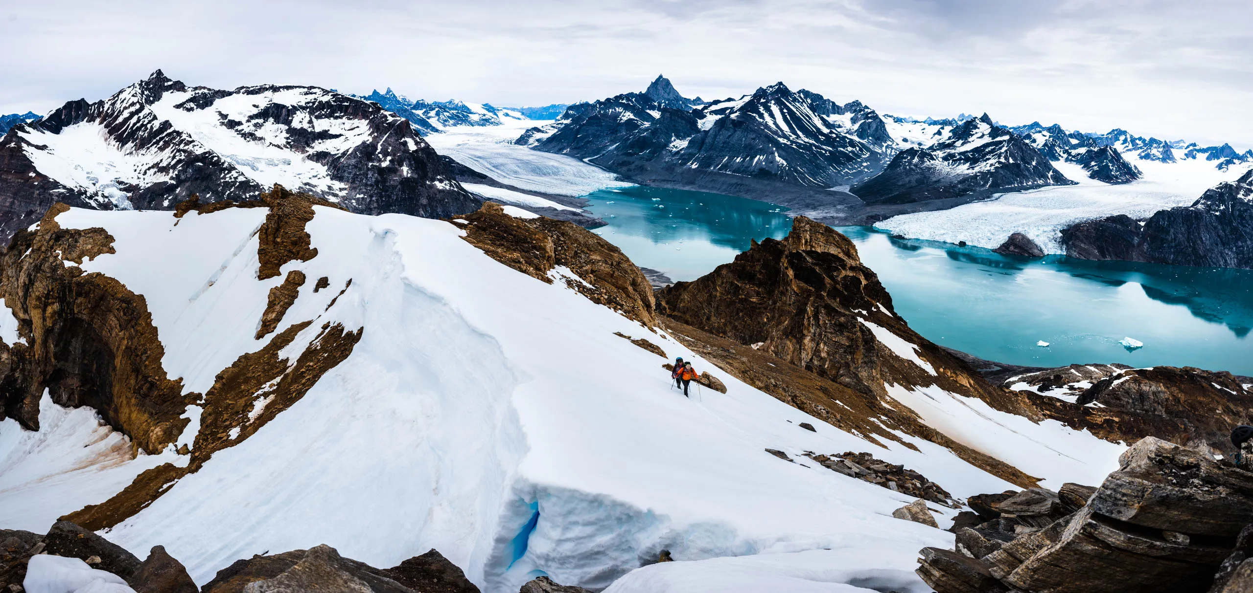 Two mountaineers traverse a snowy ridge high above a glacier-filled fjord in East Greenland