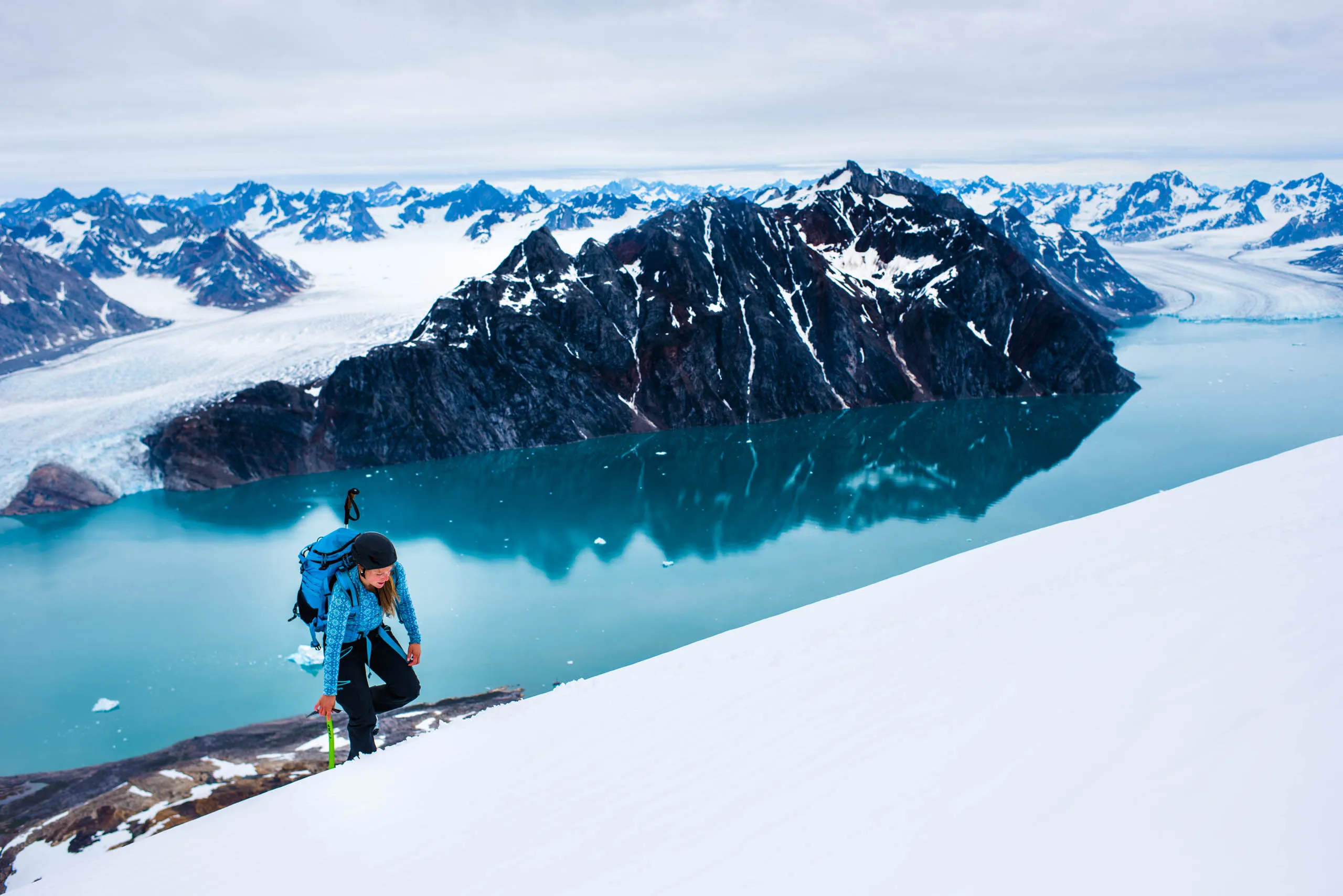 Woman climbing a snowy slope above a glacier-ringed turquoise fjord in East Greenland