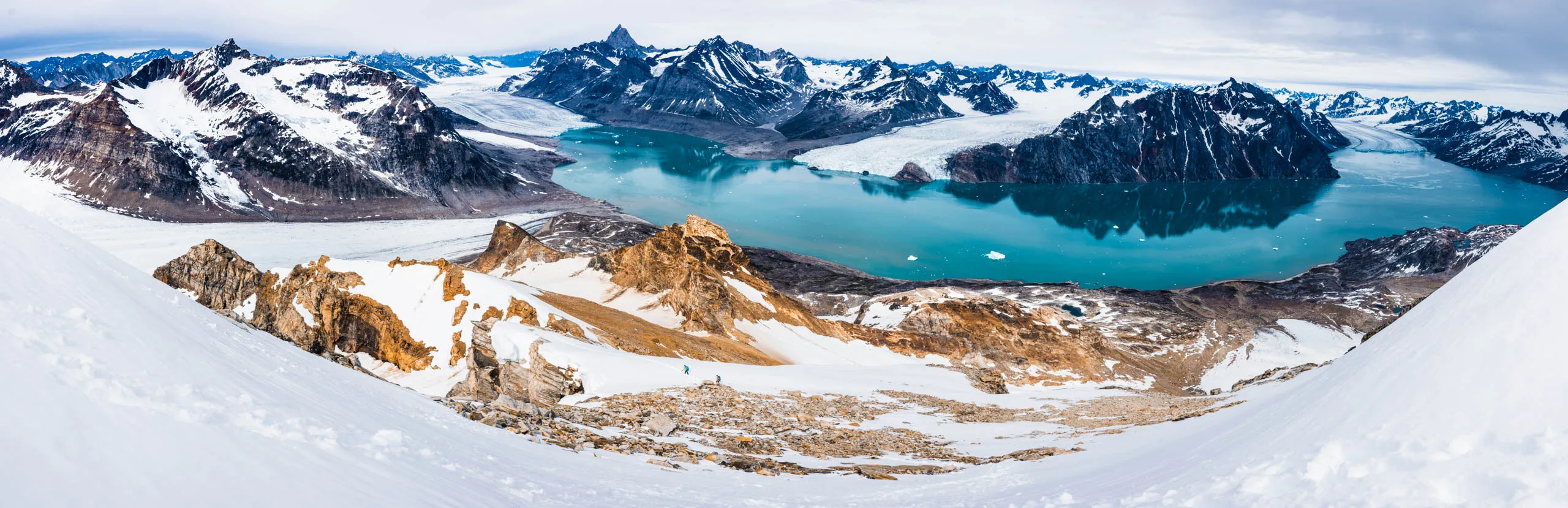 Wide panorama of two tiny hikers dwarfed by a vast snow and glacier landscape above a fjord