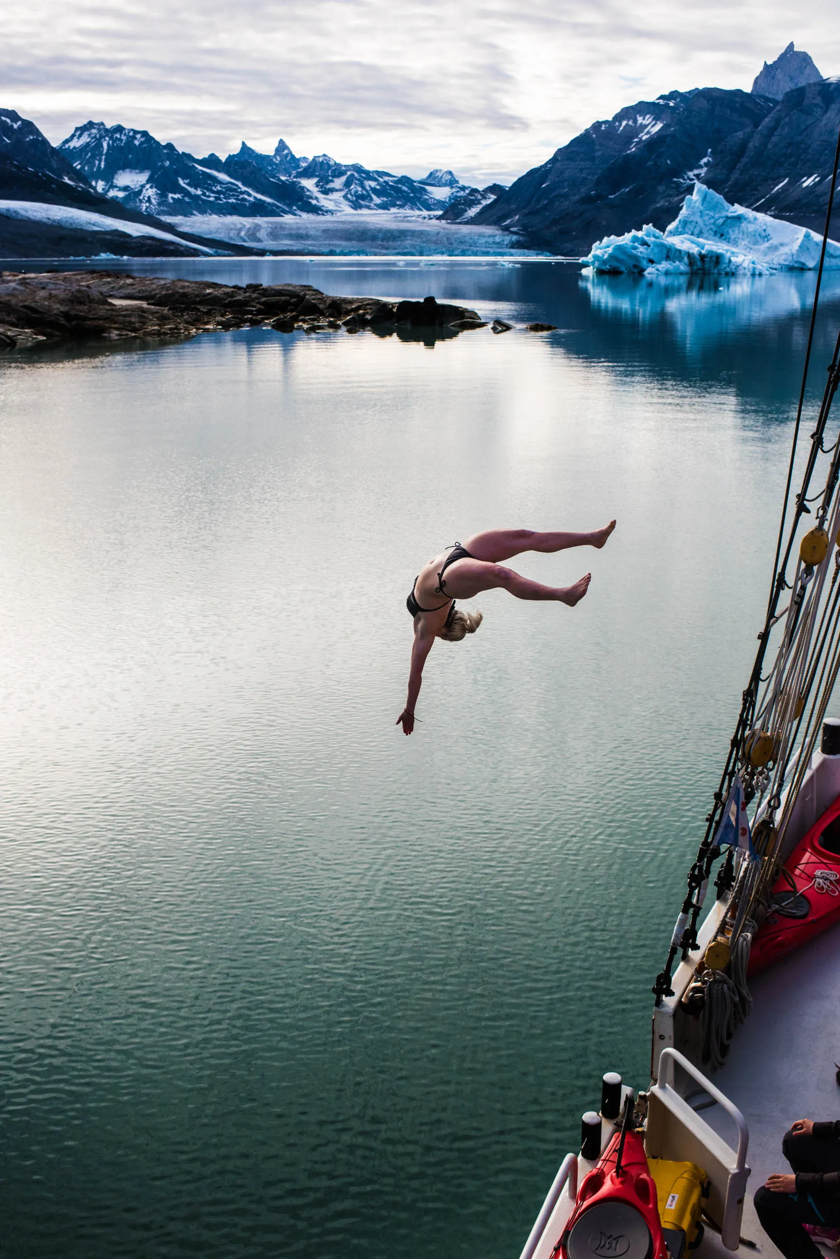 Woman diving from a sailboat into icy Arctic waters with icebergs and mountains behind her