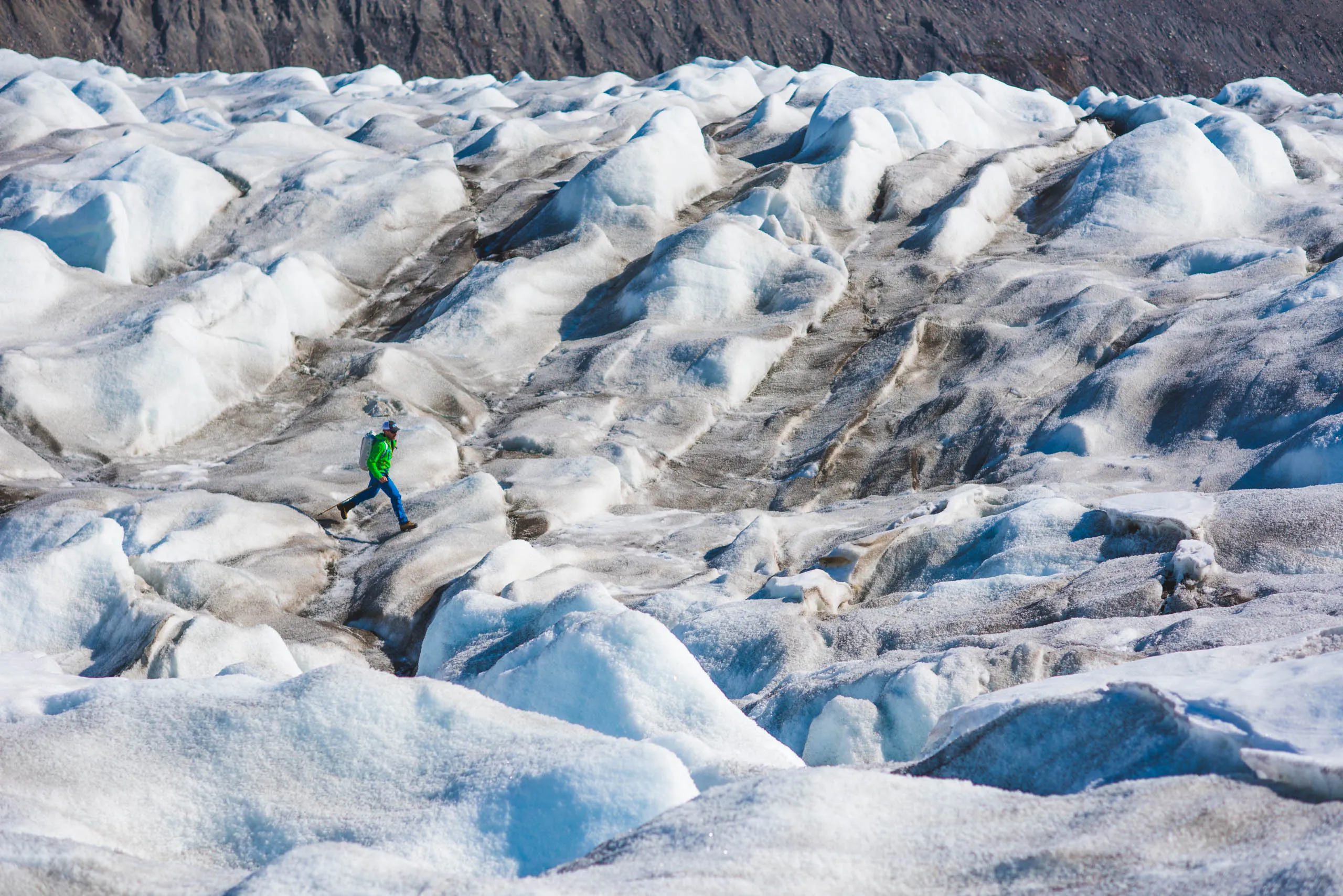Lone hiker in green jacket picking their way across a vast, crevassed glacier surface