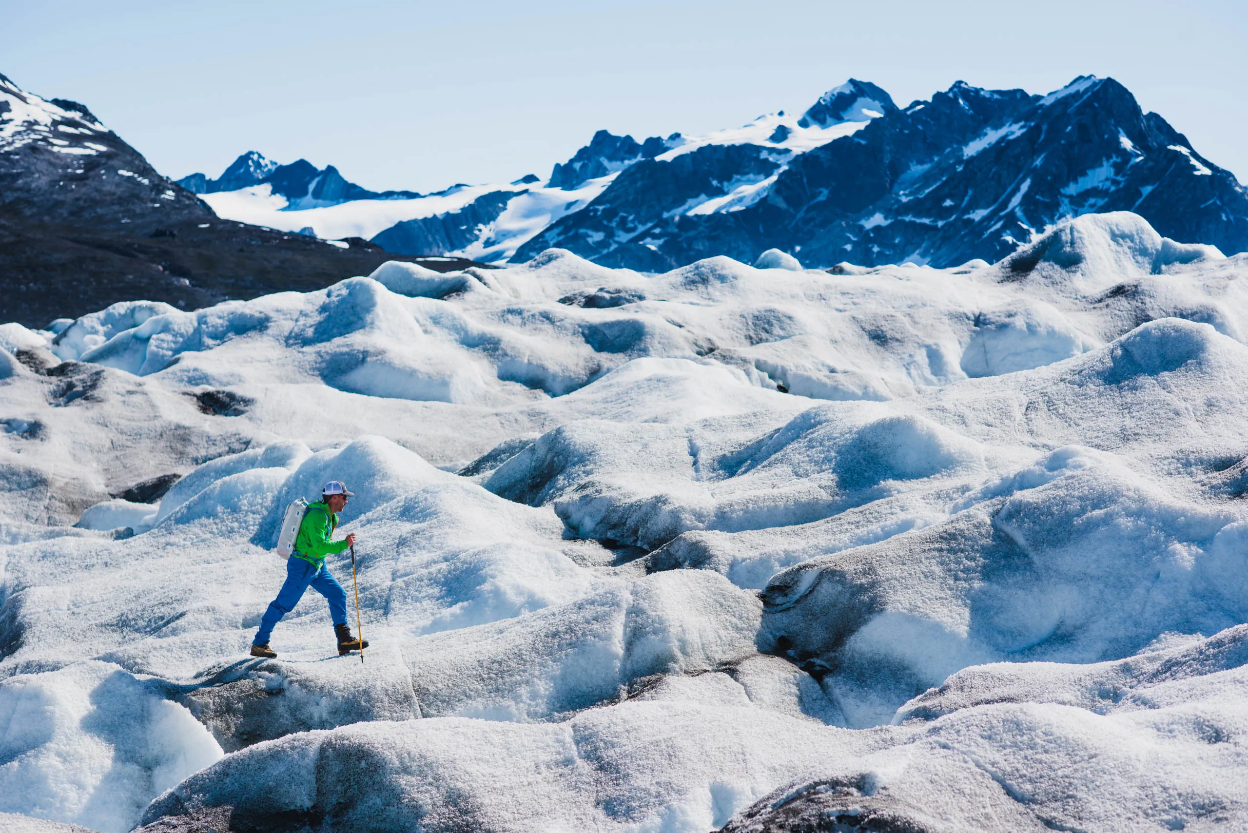 Climber with ice axe and poles traversing a sunlit glacier with jagged snow mountains behind
