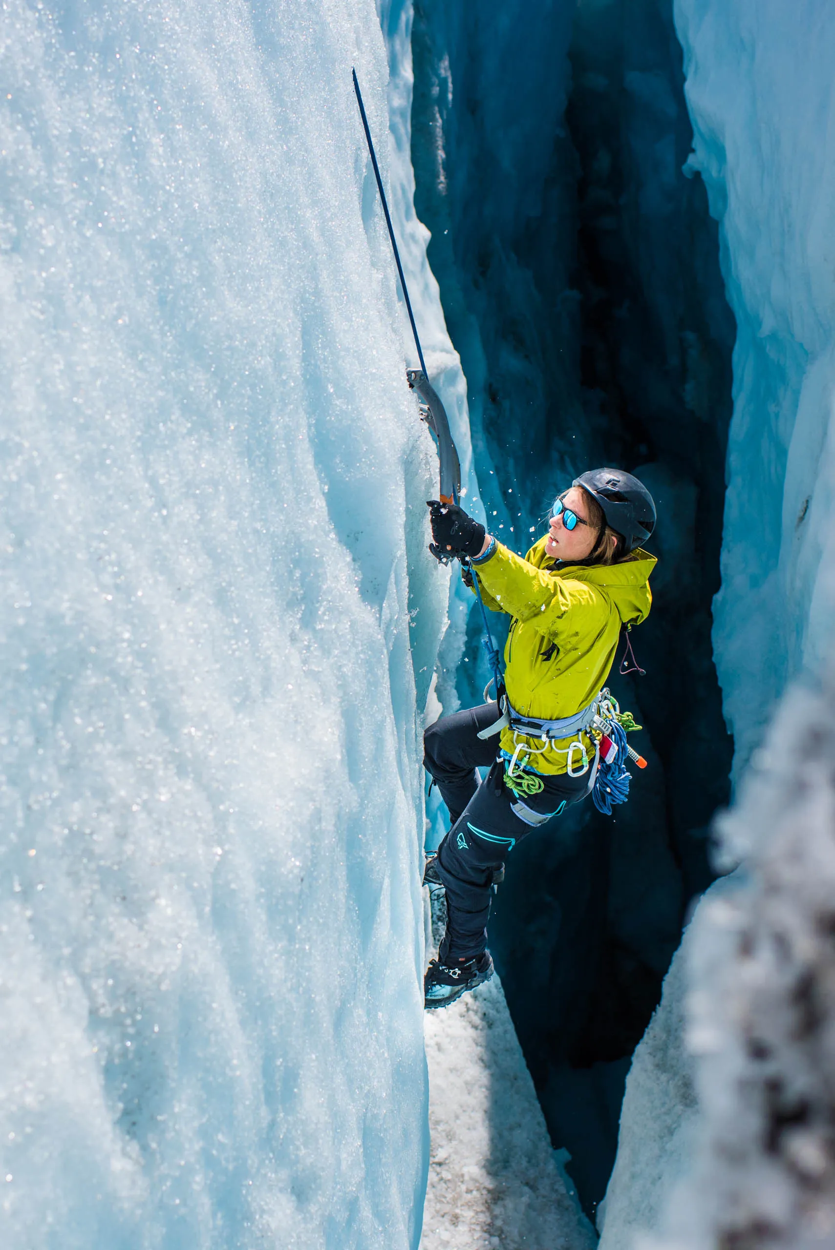 Climber in yellow jacket scaling a sheer blue glacier wall deep inside a narrow crevasse