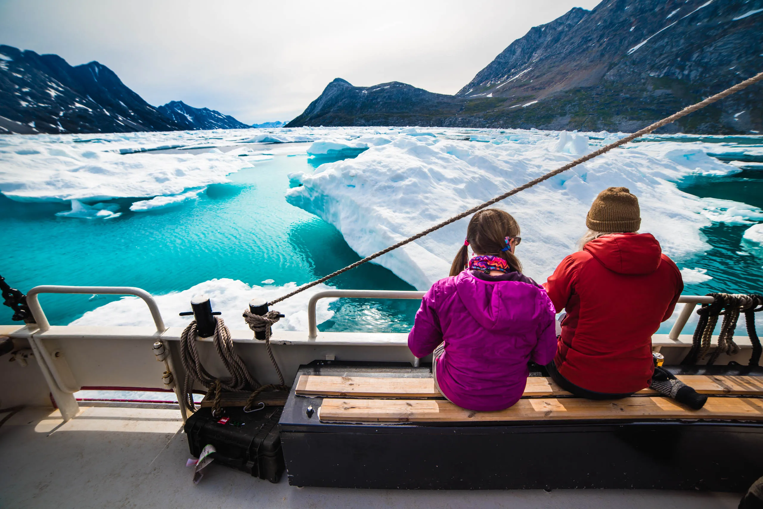 Two passengers sit on deck watching turquoise ice-filled fjord and snowy mountains