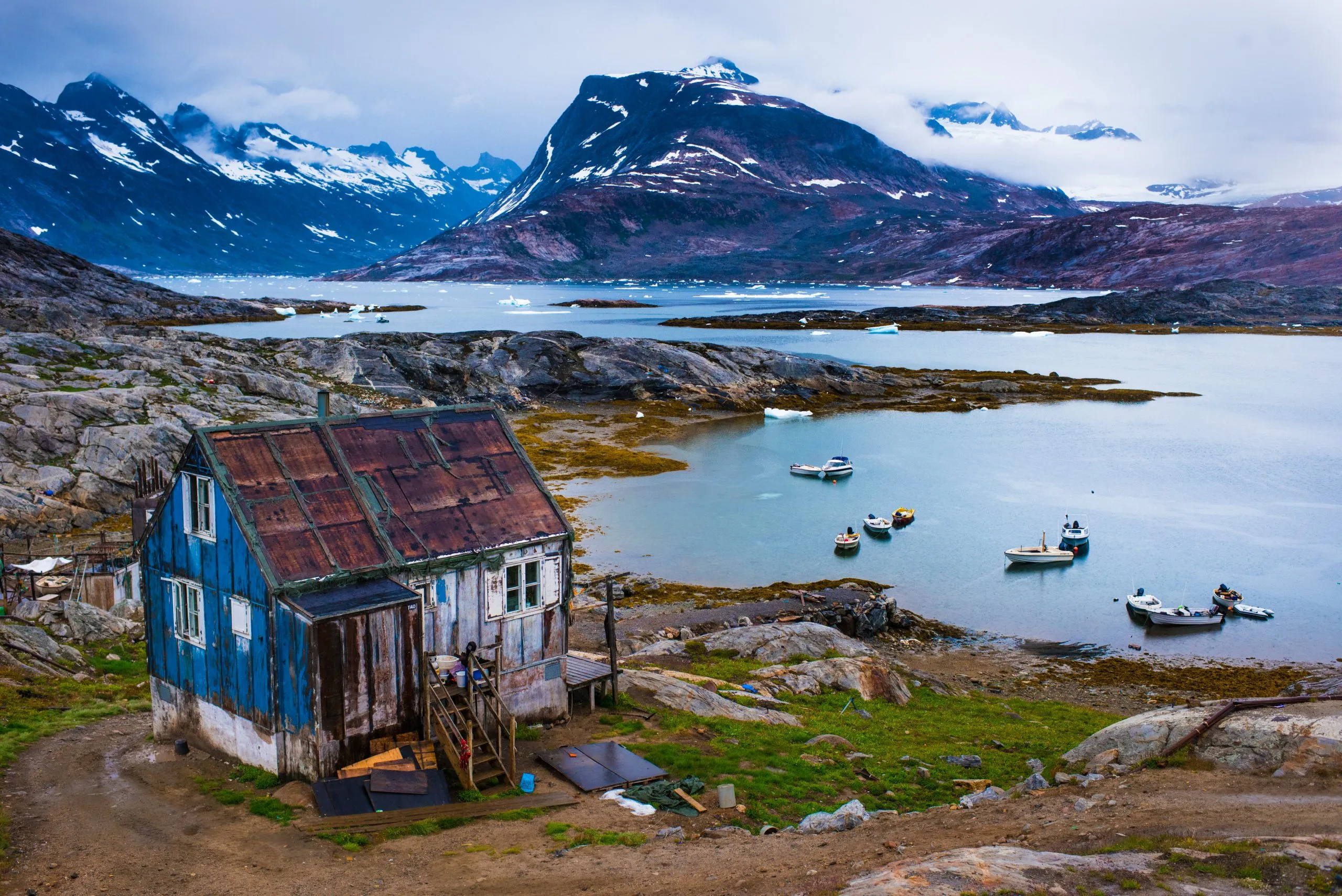 A weathered blue-and-white wooden settlement house stands on a rocky shore in front of an icy fjord and snow-capped mountains in East Greenland.