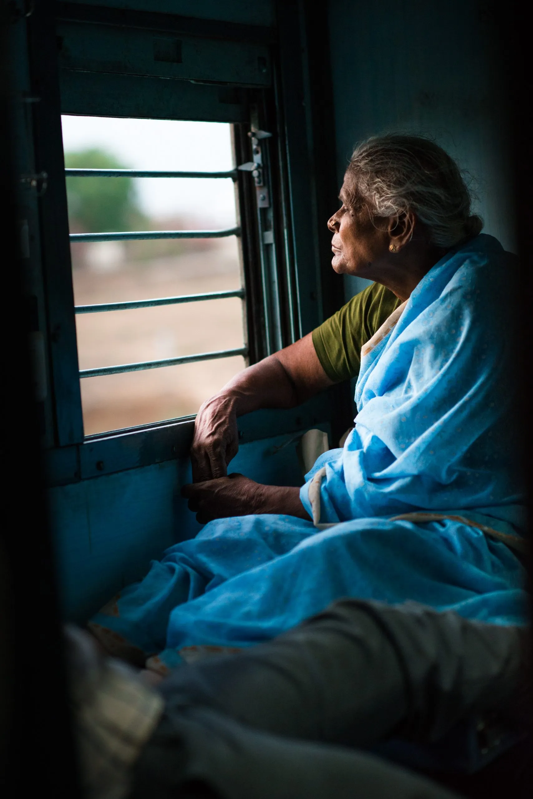 An older Indian woman in a blue sari leans against the barred window of a moving Indian Railways train carriage, gazing out at the passing countryside.