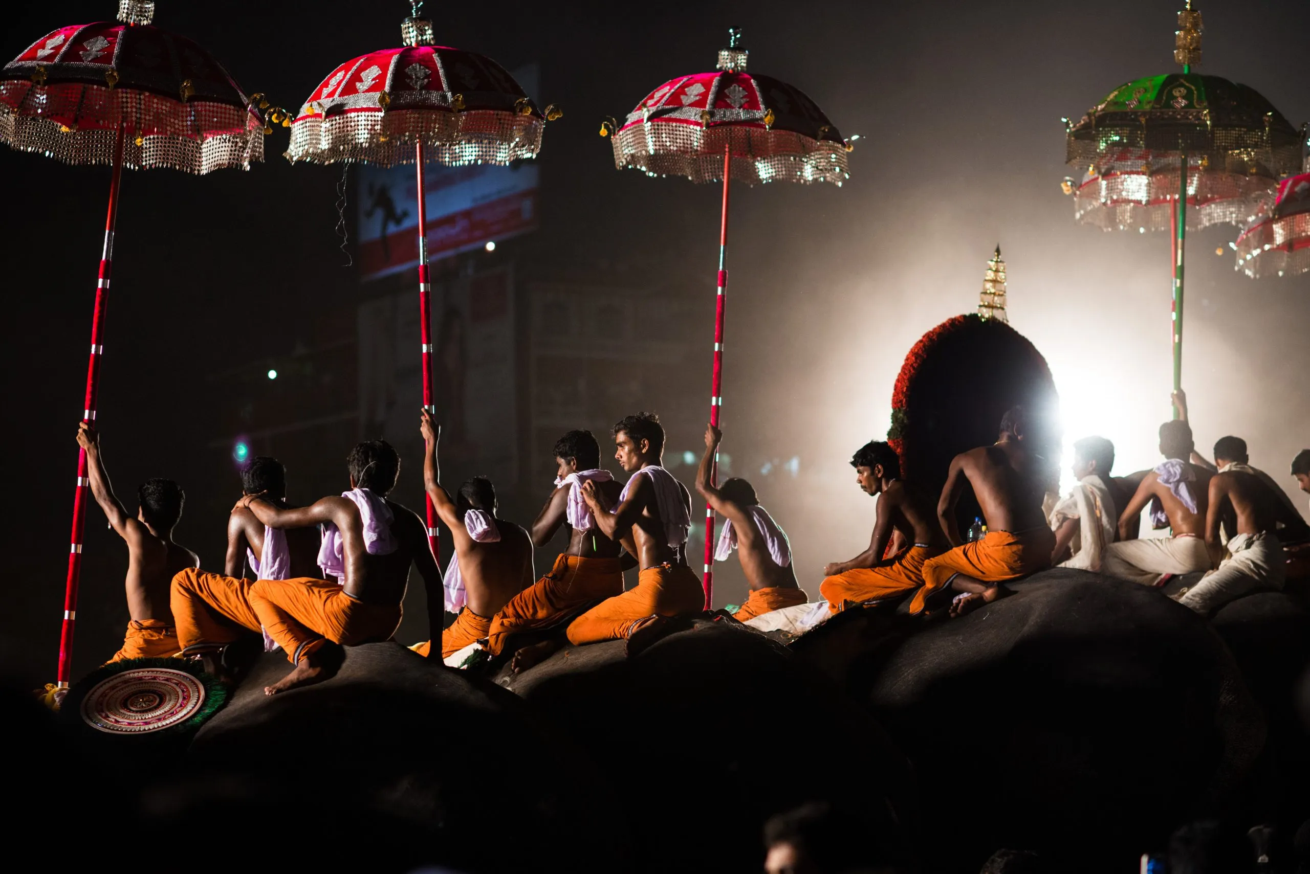 Men in saffron dhotis sit on caparisoned elephants holding tall sequinned ceremonial parasols at the Thrissur Pooram festival in Kerala, India.