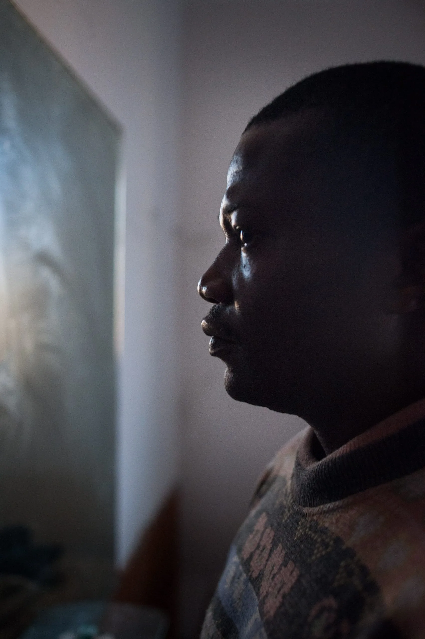 A young man's profile in dim window-light beside a polished metal surface, inside the Rosarno warehouse.