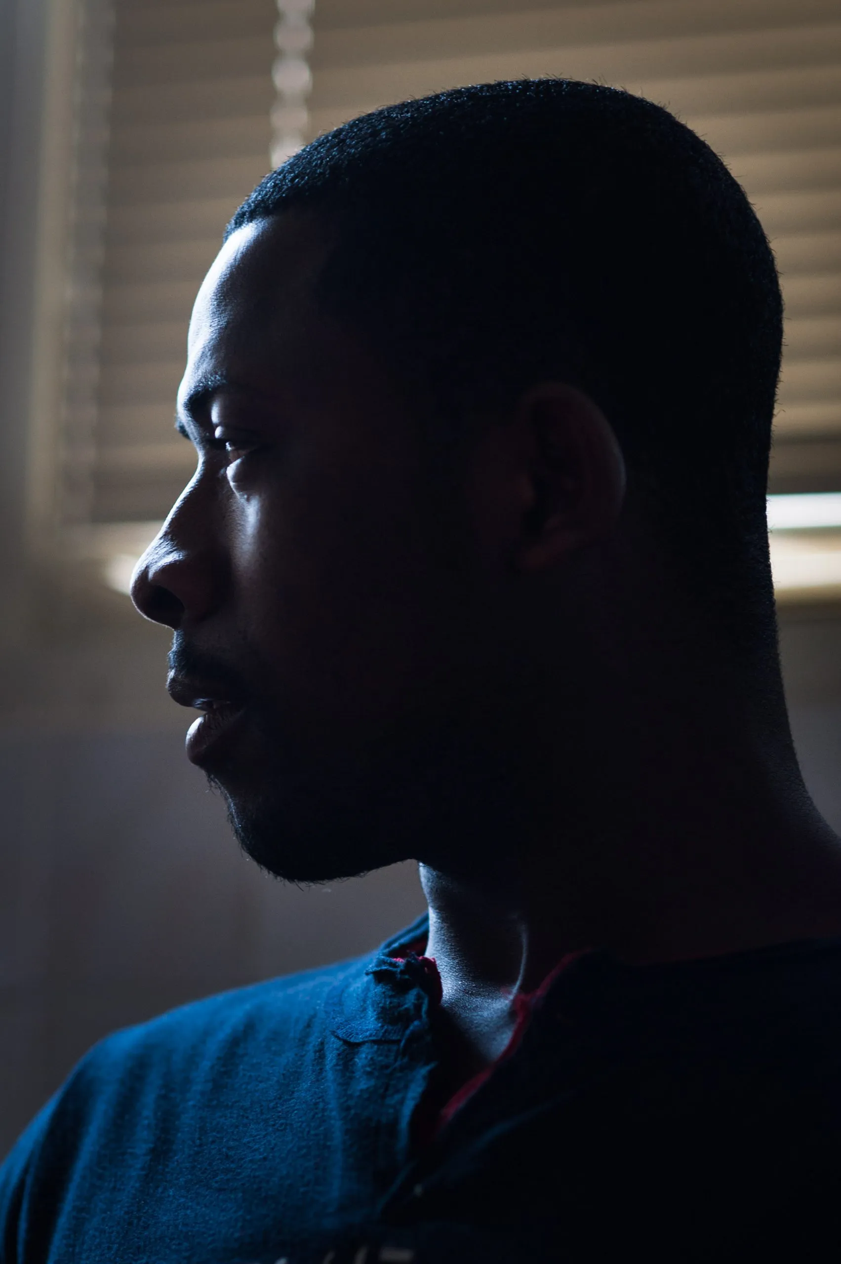 A man's profile in low light, beside a window with closed venetian blinds, inside an abandoned warehouse in Rosarno, Italy.
