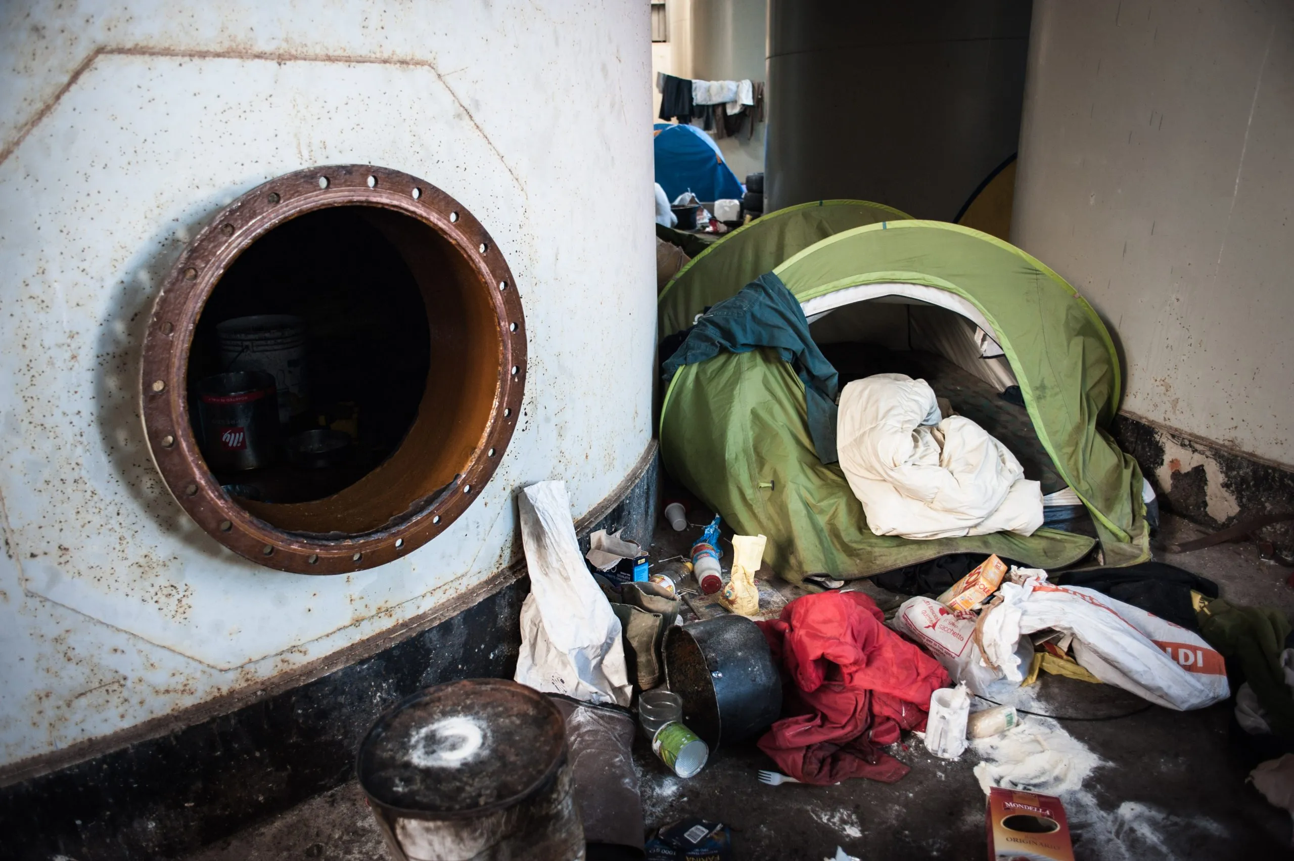 View through a steel pipe opening of a tent and scattered belongings on the warehouse floor in Rosarno, Italy.