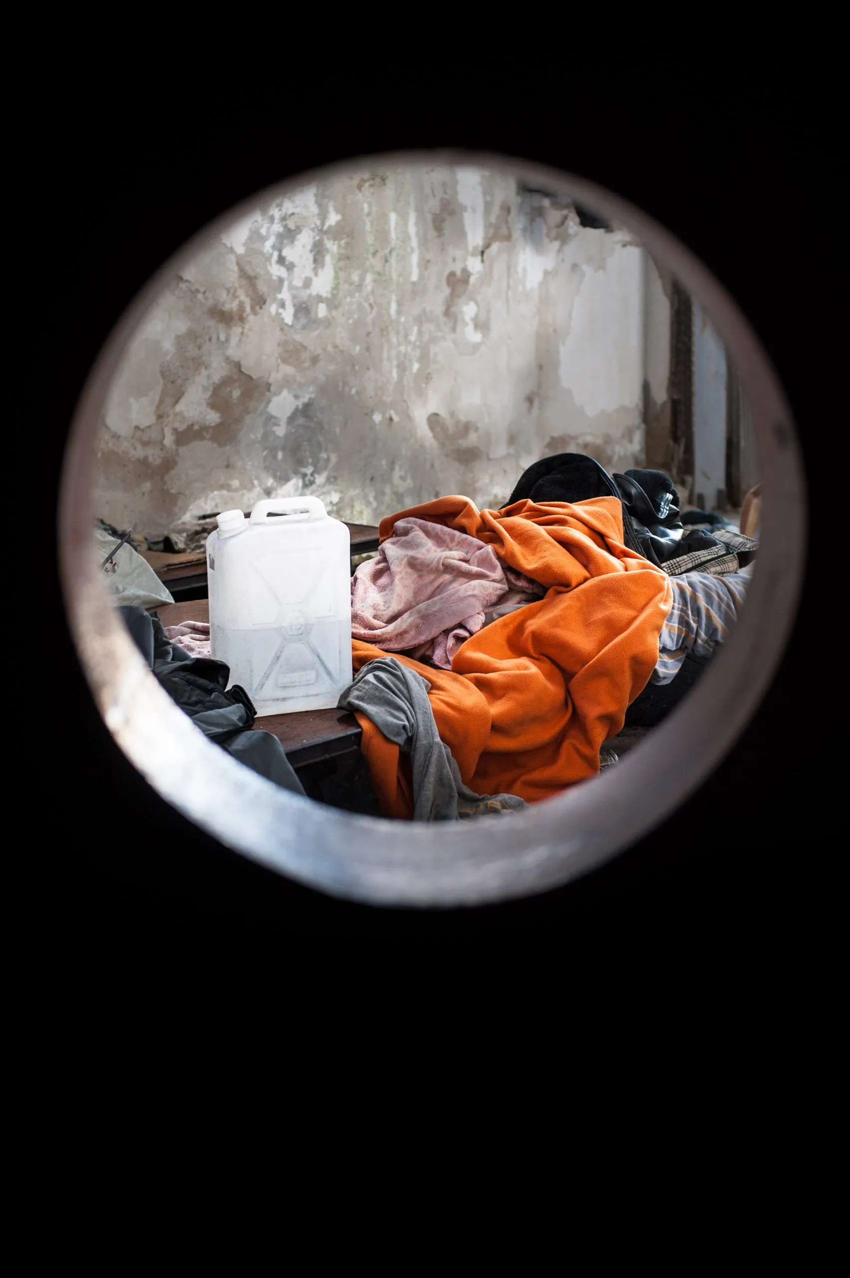 View through a steel pipe opening of bedding, a water canister and peeling walls inside the Rosarno warehouse.