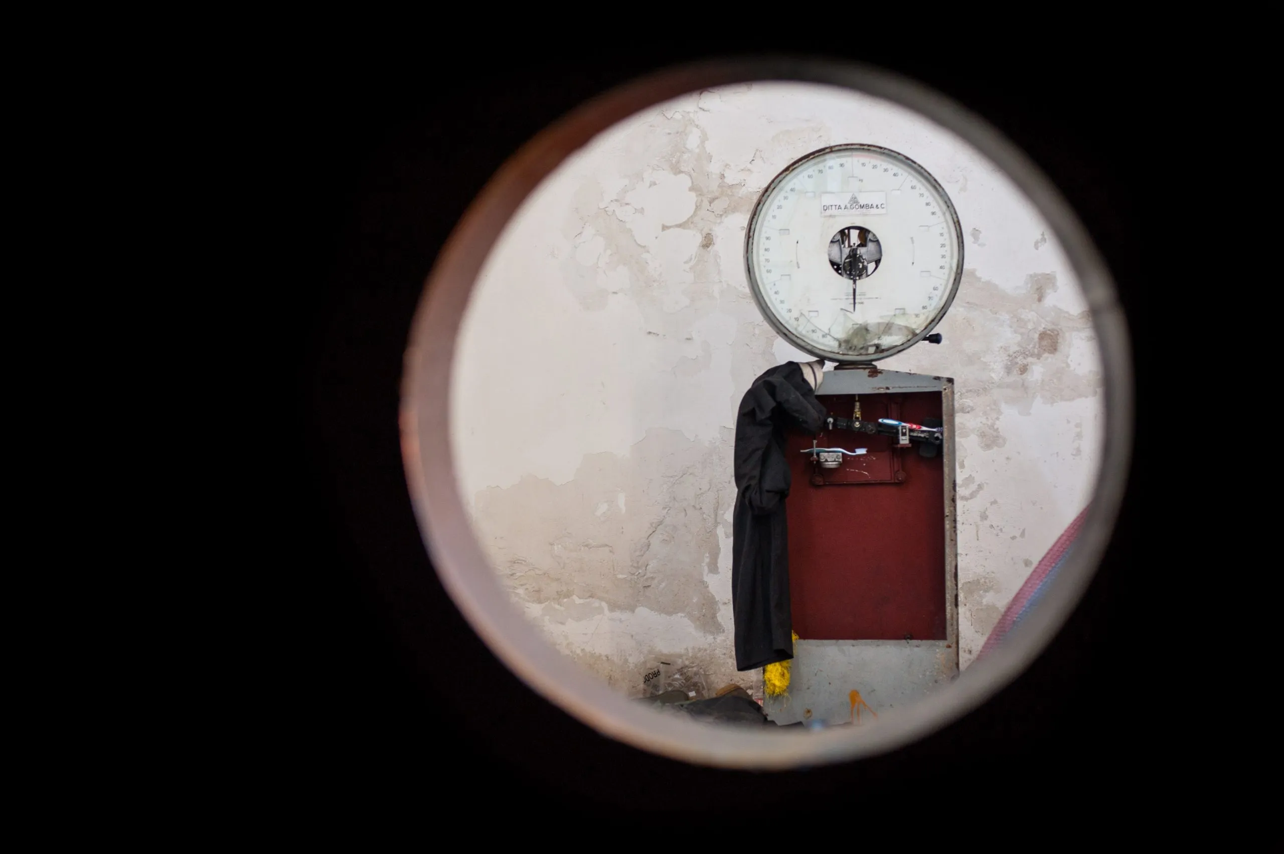 An old Italian weighing scale on a peeling wall, seen through a steel pipe opening in the abandoned warehouse.