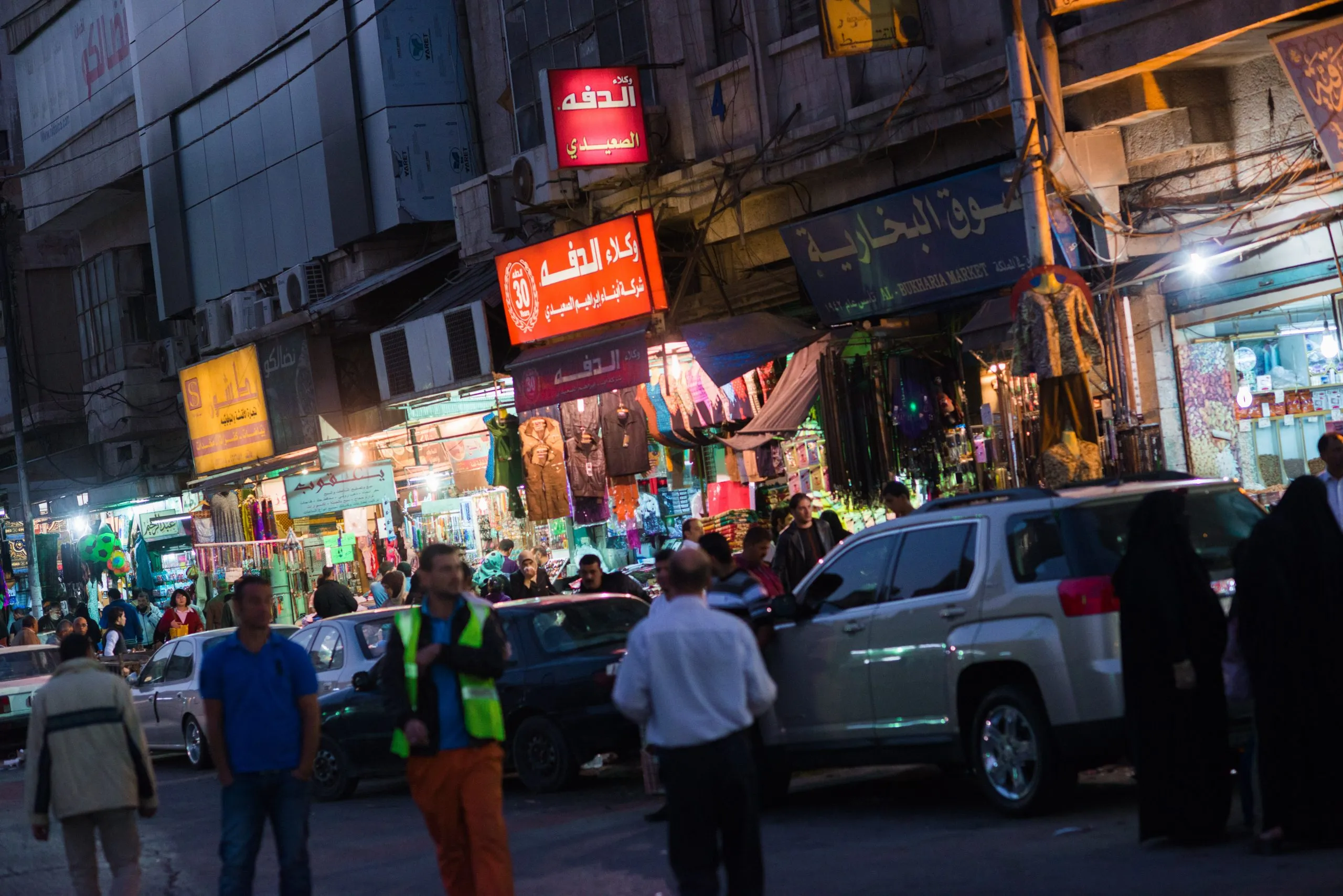 A bustling night market in central Amman, Jordan, with neon shop signs in Arabic and people moving between cars.