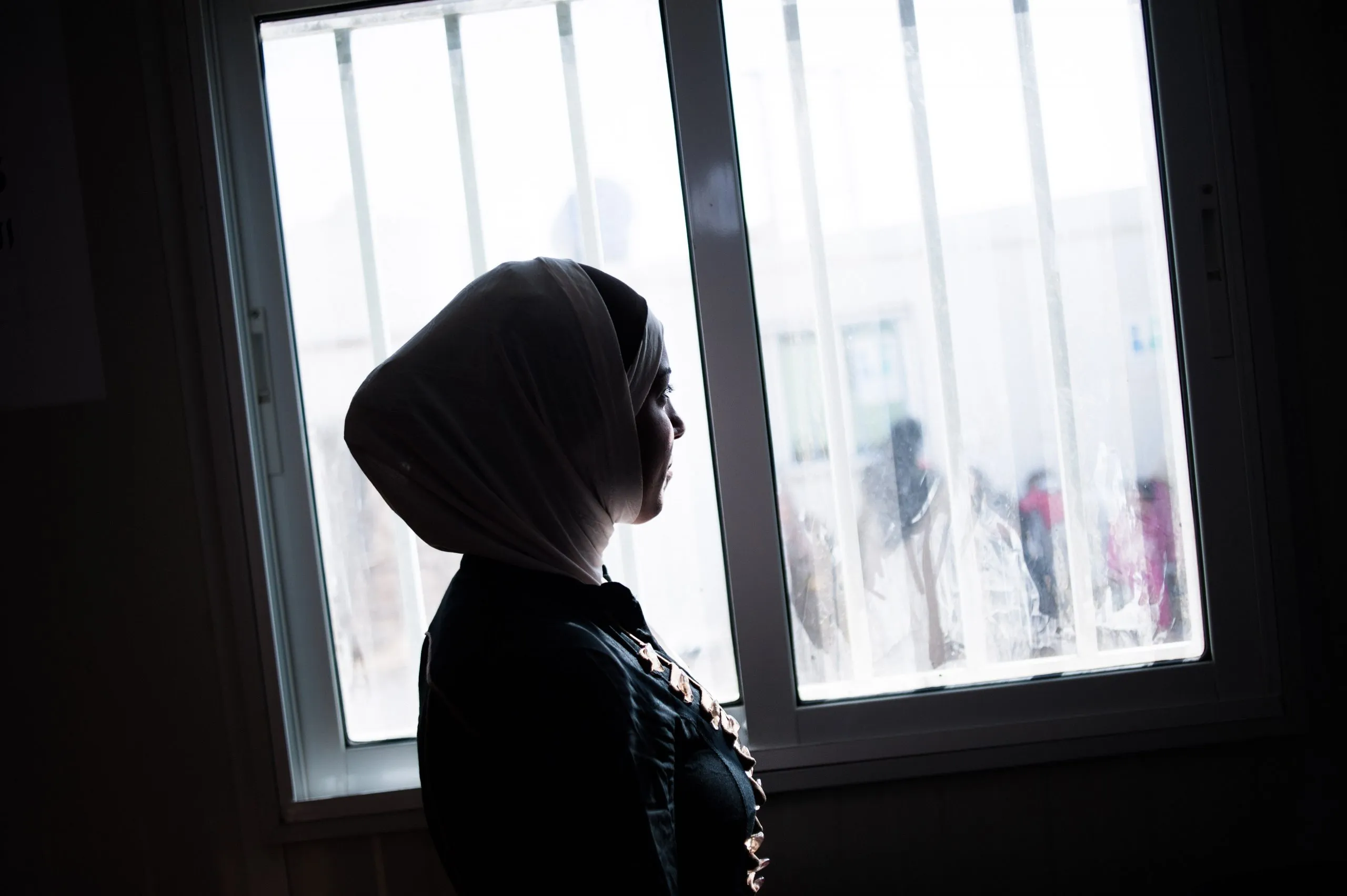 A woman in a headscarf stands in silhouette at a barred window, looking out at an indistinct courtyard in Jordan.