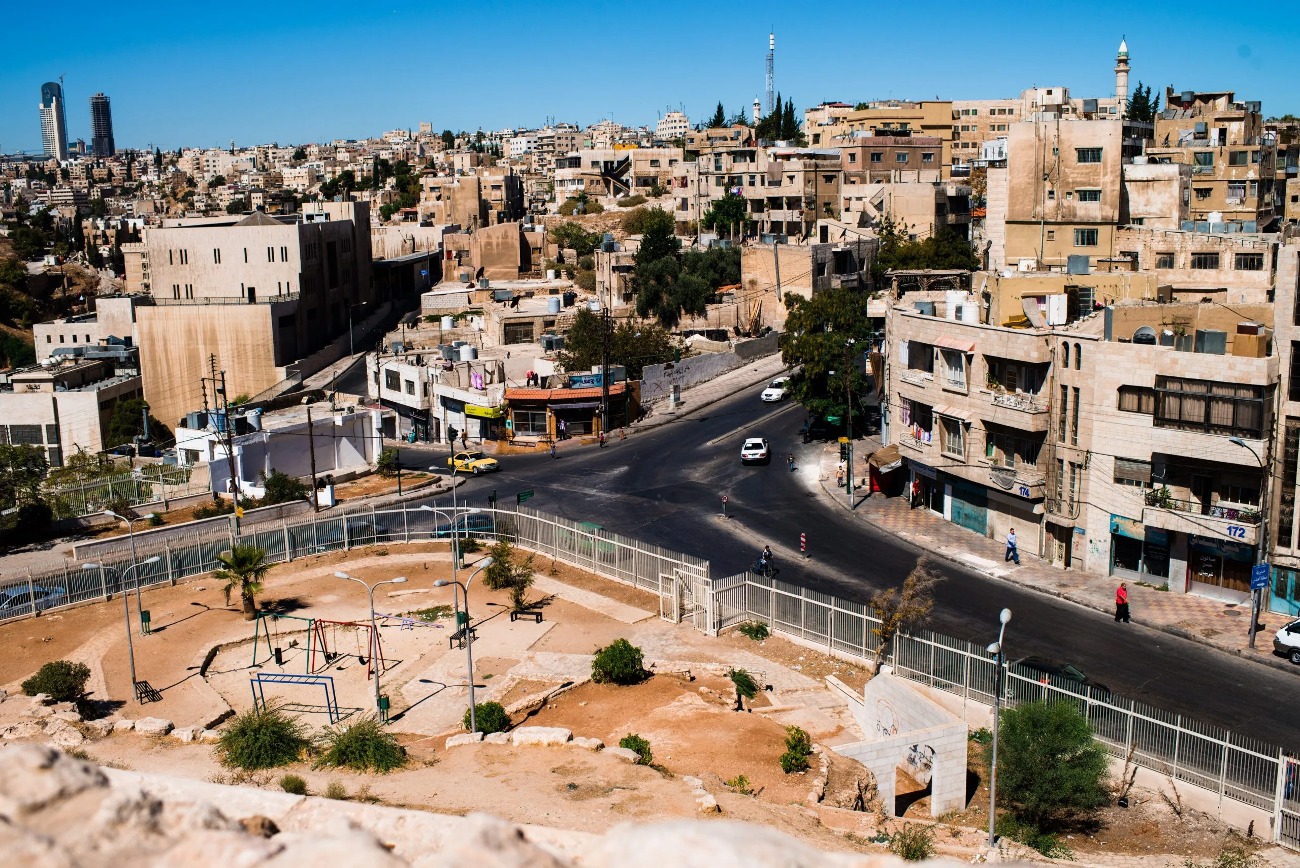 An empty playground beside a steep road in Amman, Jordan, against the city's distinctive yellow stone buildings.