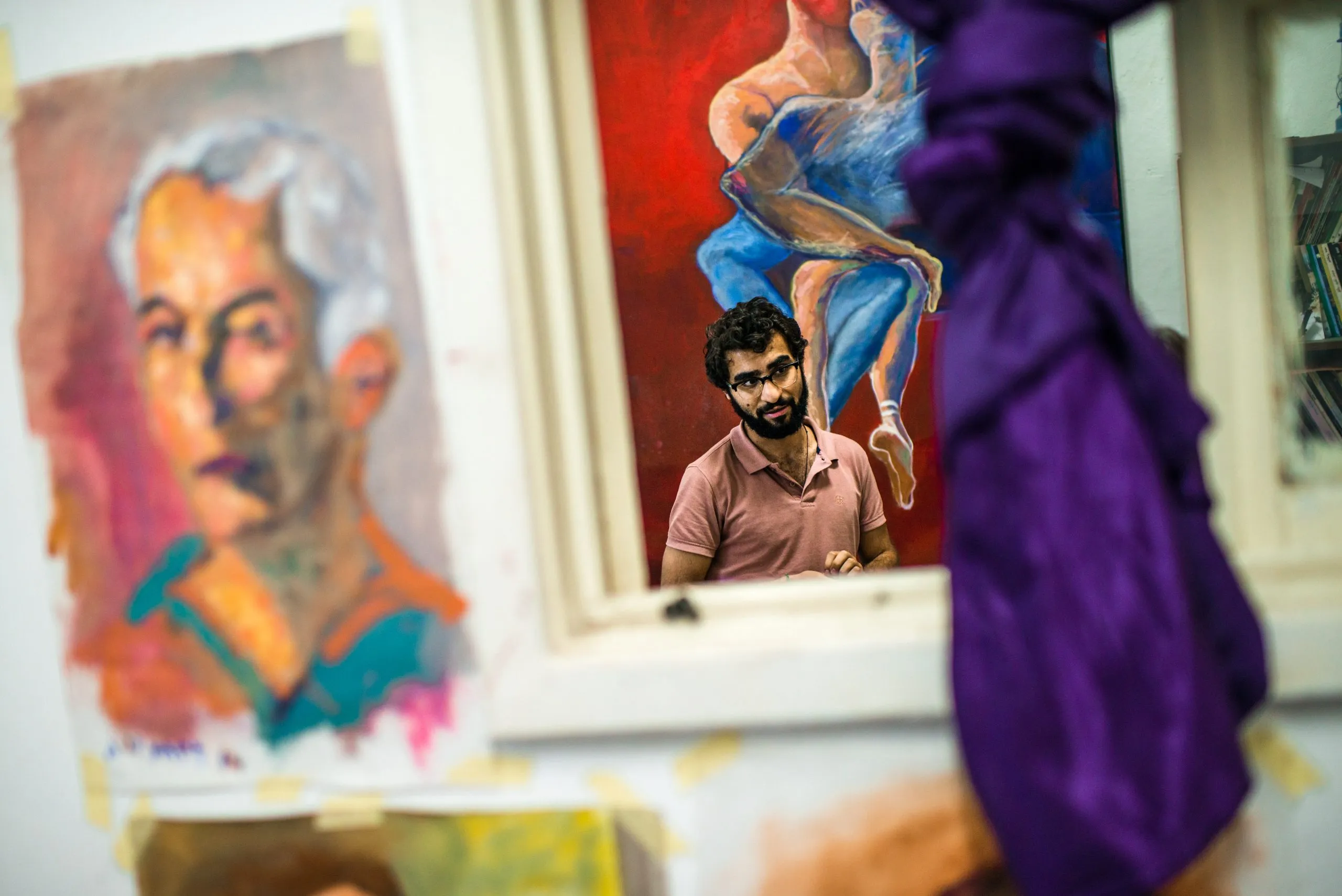 A young artist stands among his oil paintings inside a small studio in Amman, framed by hanging fabric.