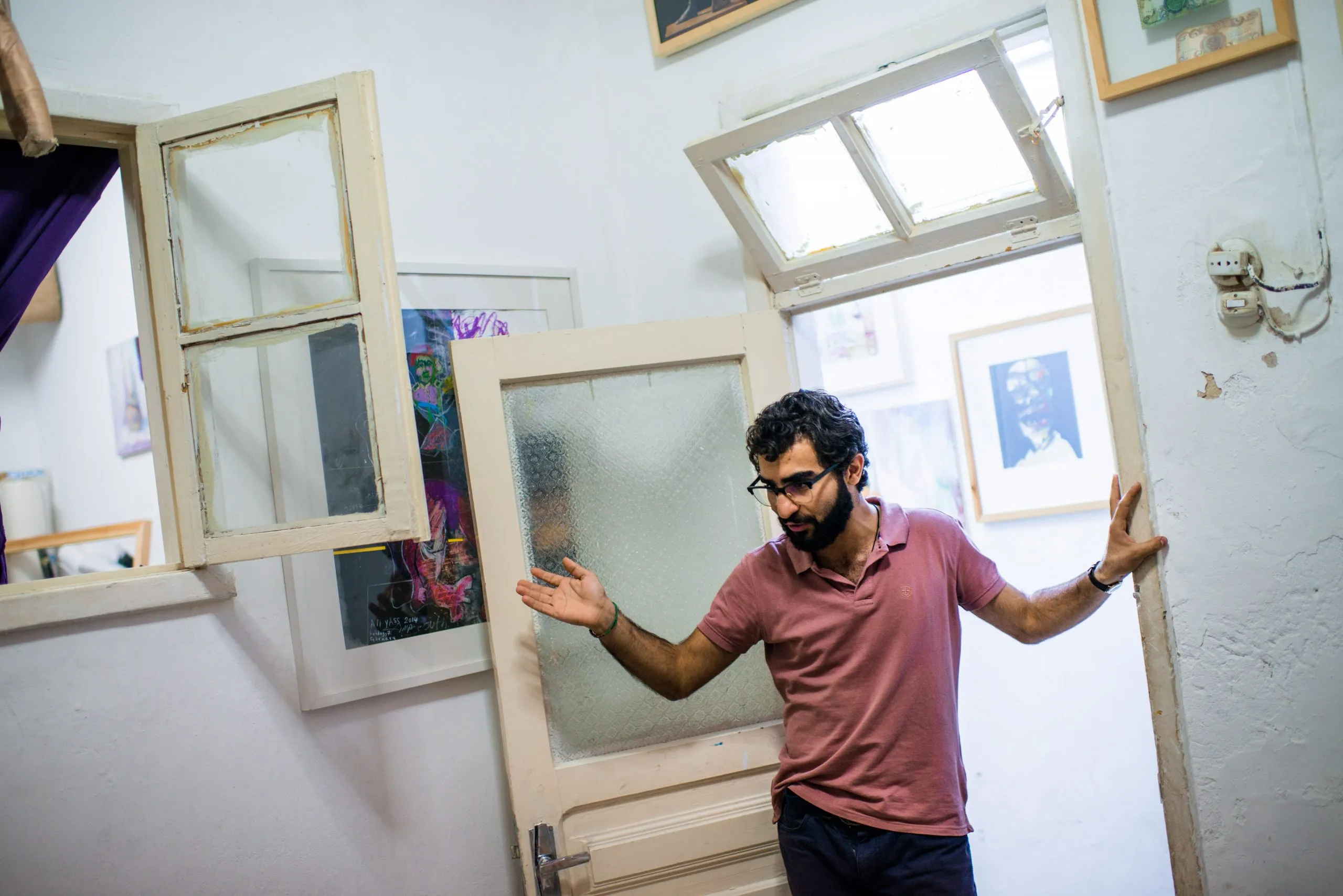 A young artist gestures at the door of his small art studio in Amman, walls hung with framed sketches.