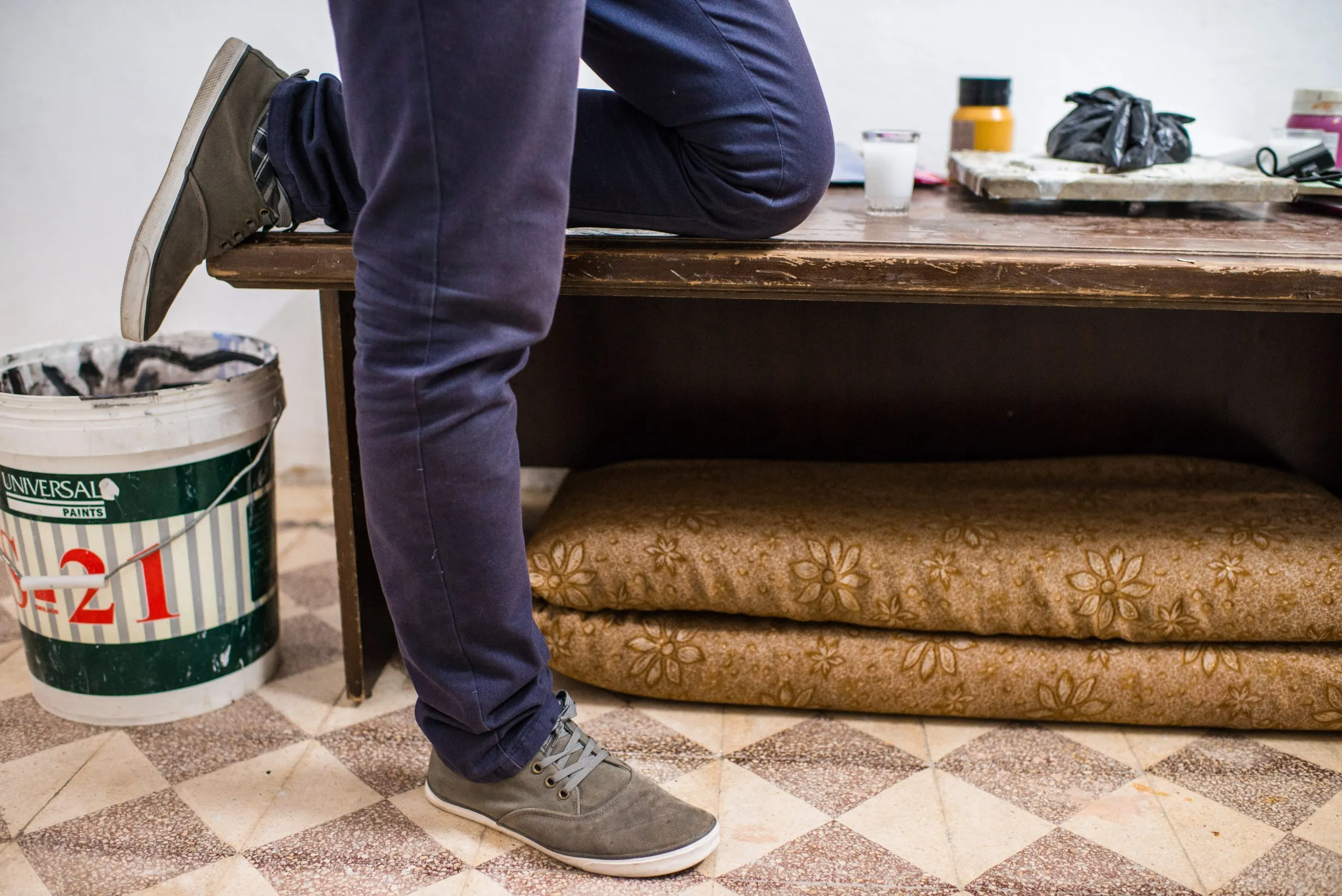 A person sits cross-legged on a worn wooden bench beside paint buckets in an art space in Amman.