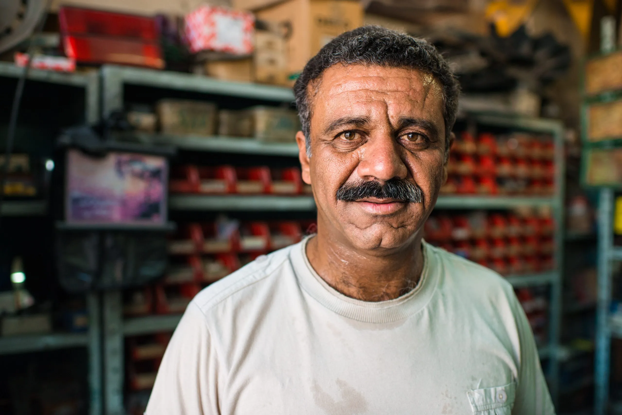 A mechanic in a white t-shirt stands among shelves of car parts in his small repair shop in Amman.