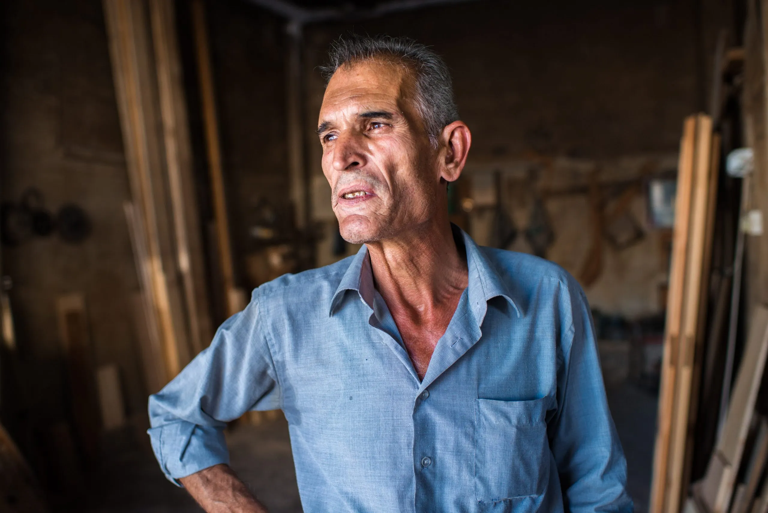 An older carpenter in a blue shirt stands in his workshop in Amman, Jordan.