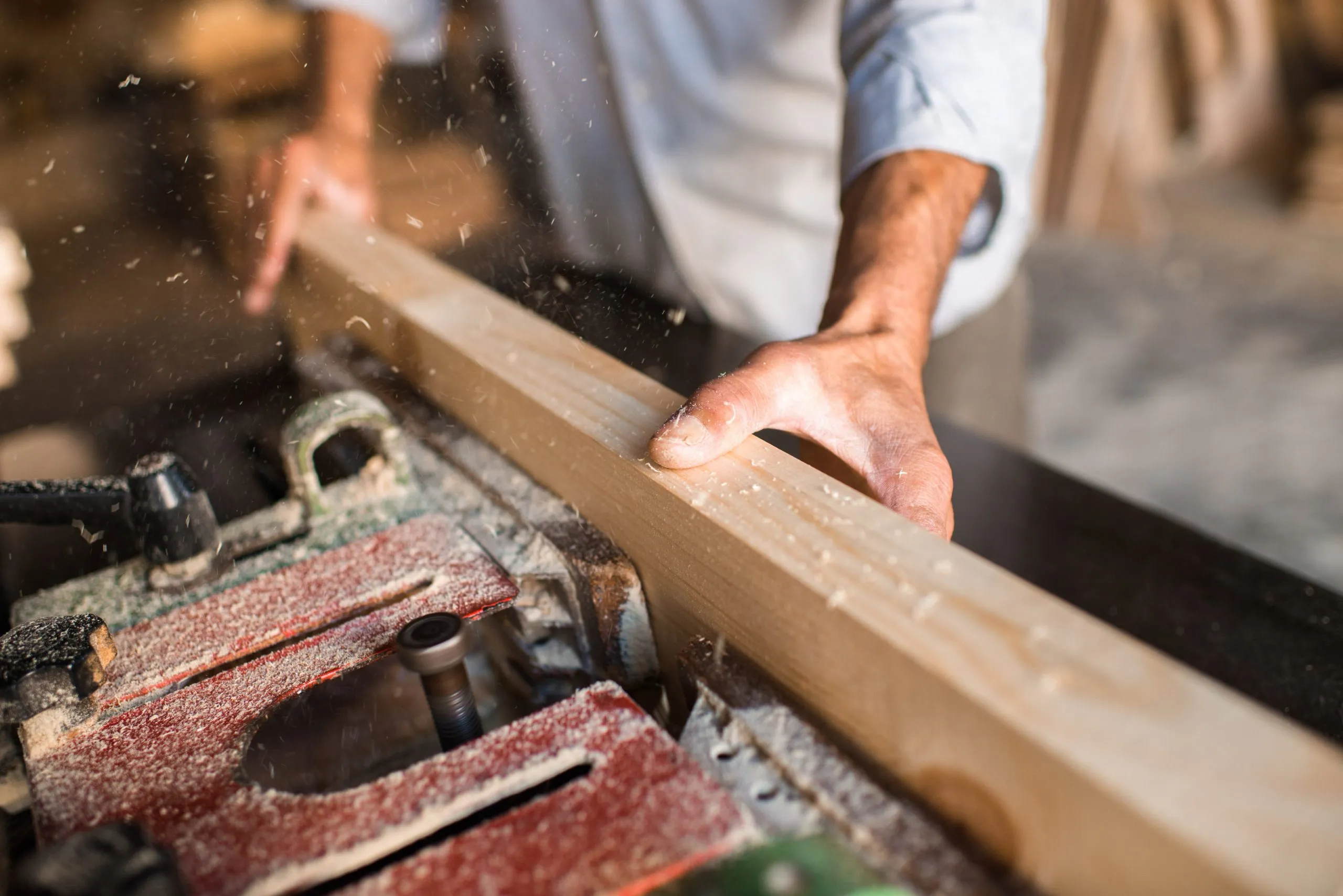 Close view of a carpenter's hands steadying a length of wood through a planer, sawdust flying, in Amman.