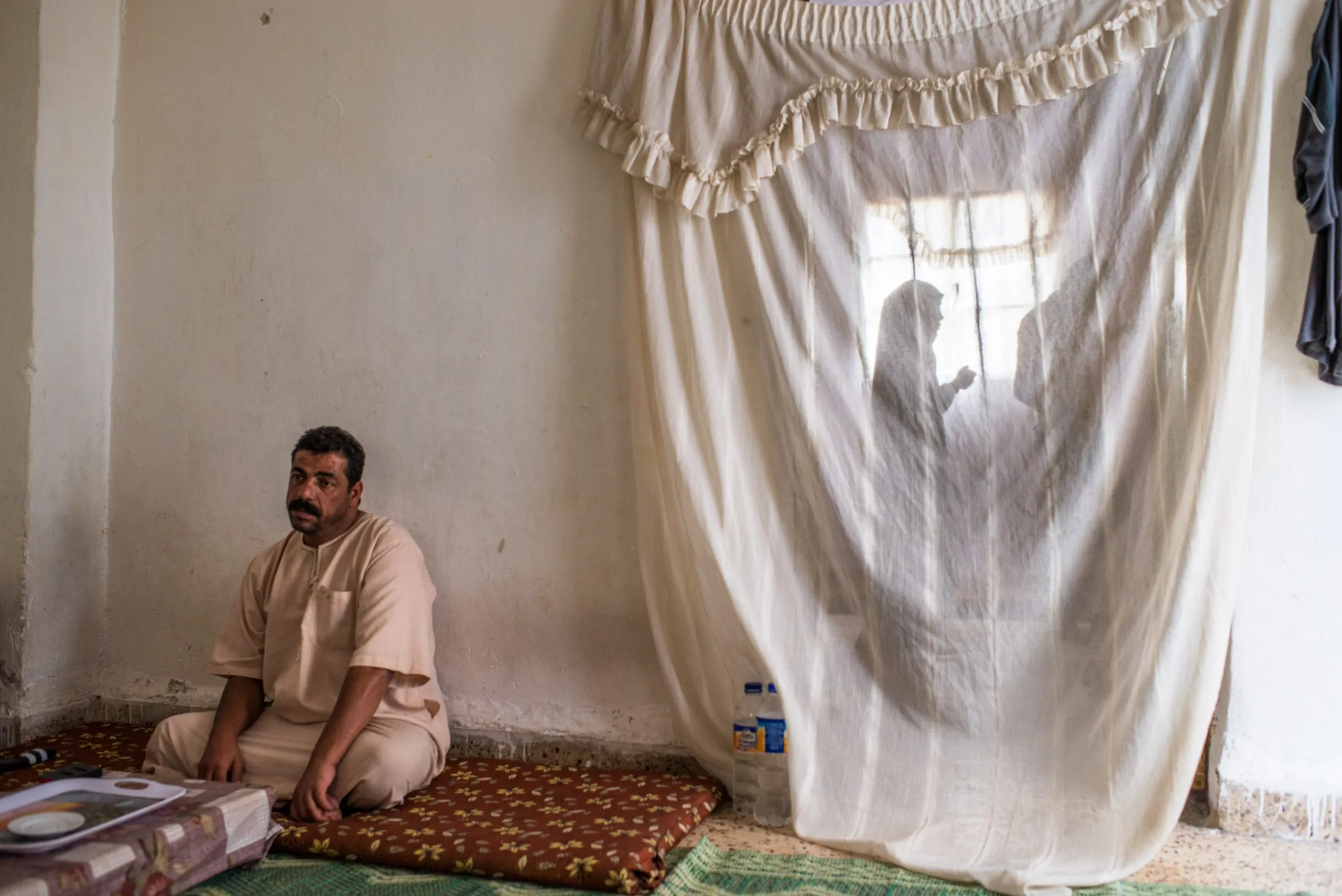 A man sits on a patterned rug at home as a woman moves behind a thin lace curtain, in Jordan.