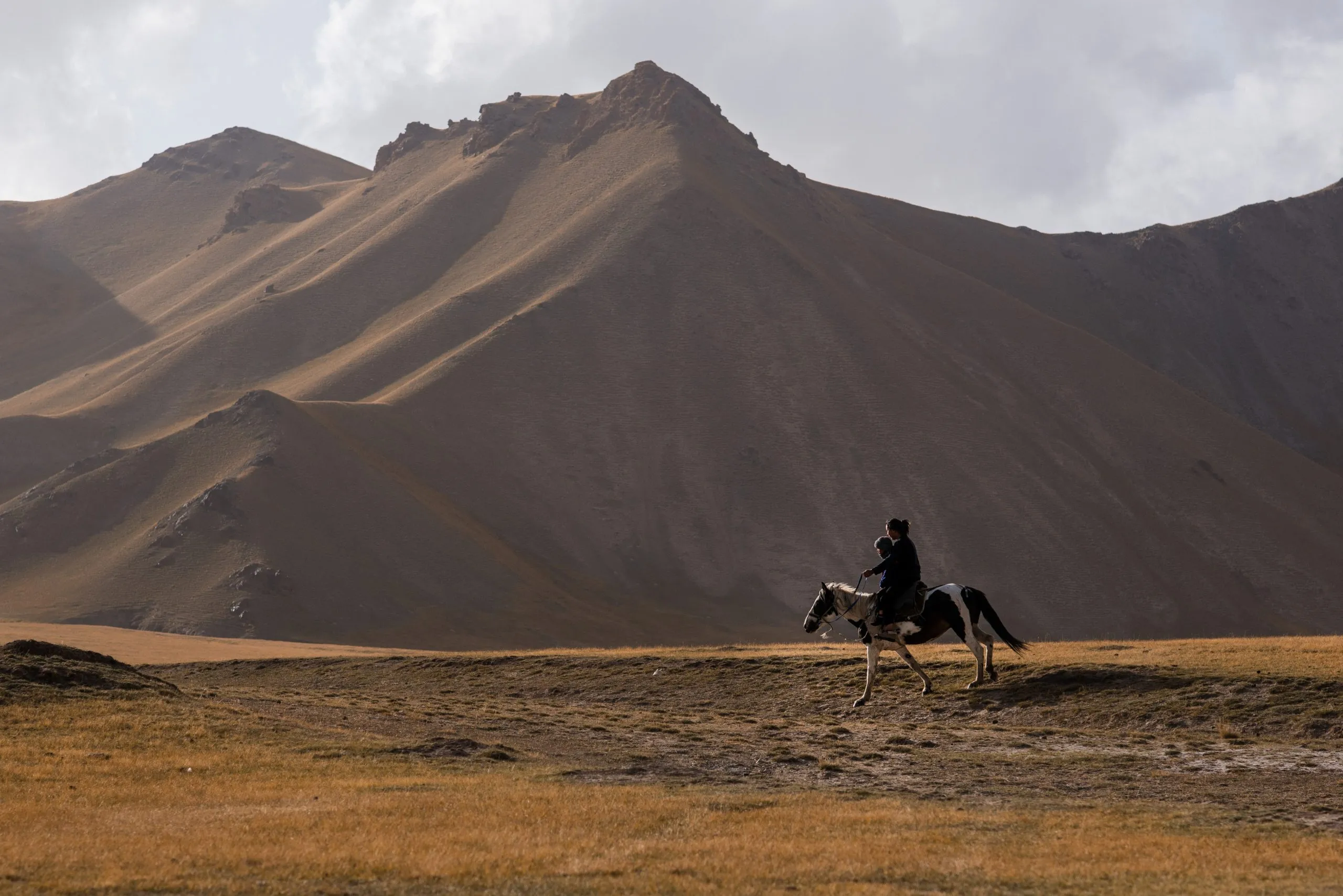 A horseback rider with a child crosses a wide grassy plain at the foot of dramatic eroded peaks in the Kyrgyz mountains.