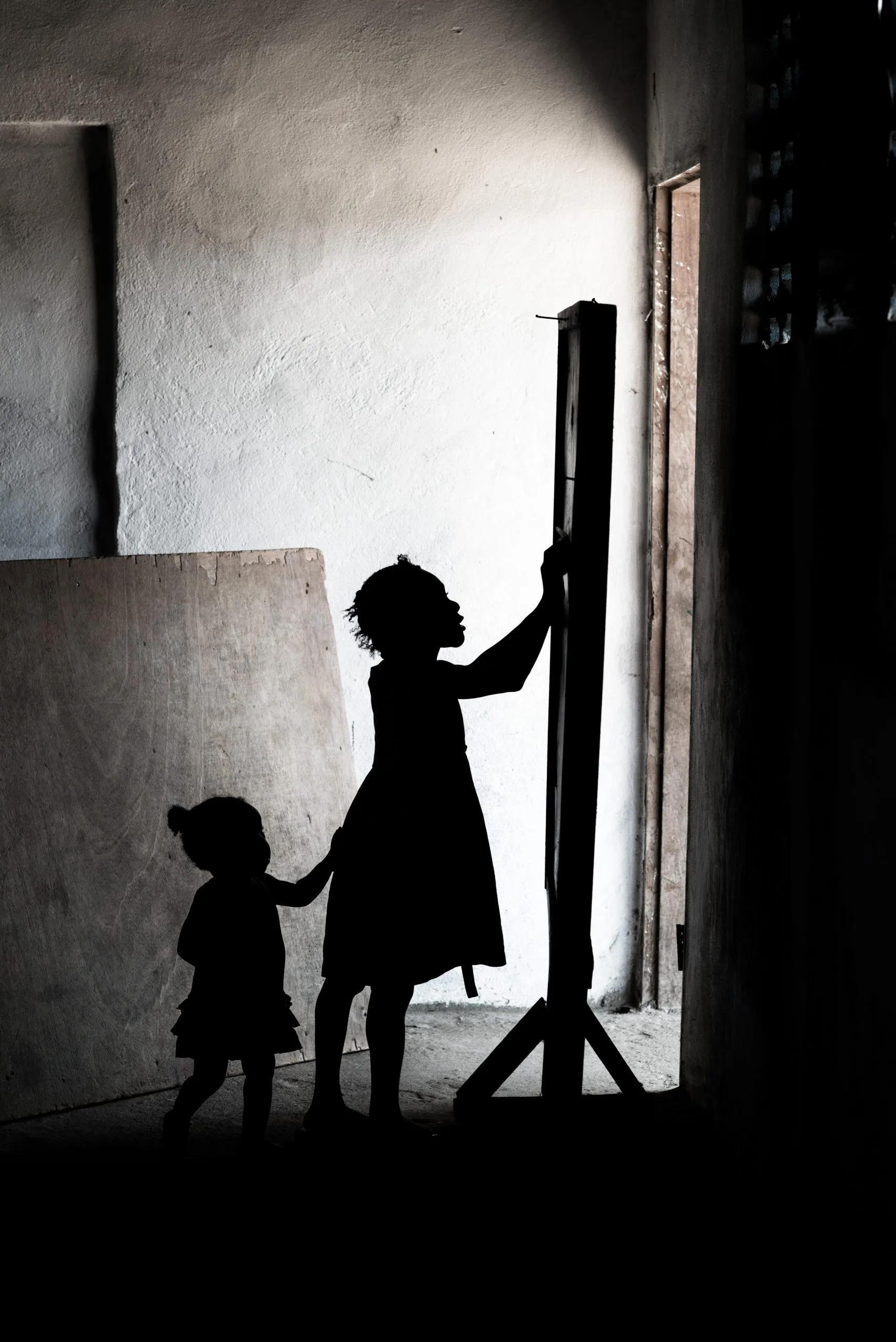 Silhouettes of two children in a sunlit doorway in Liberia, the older girl holding a toddler's hand against a wooden frame.