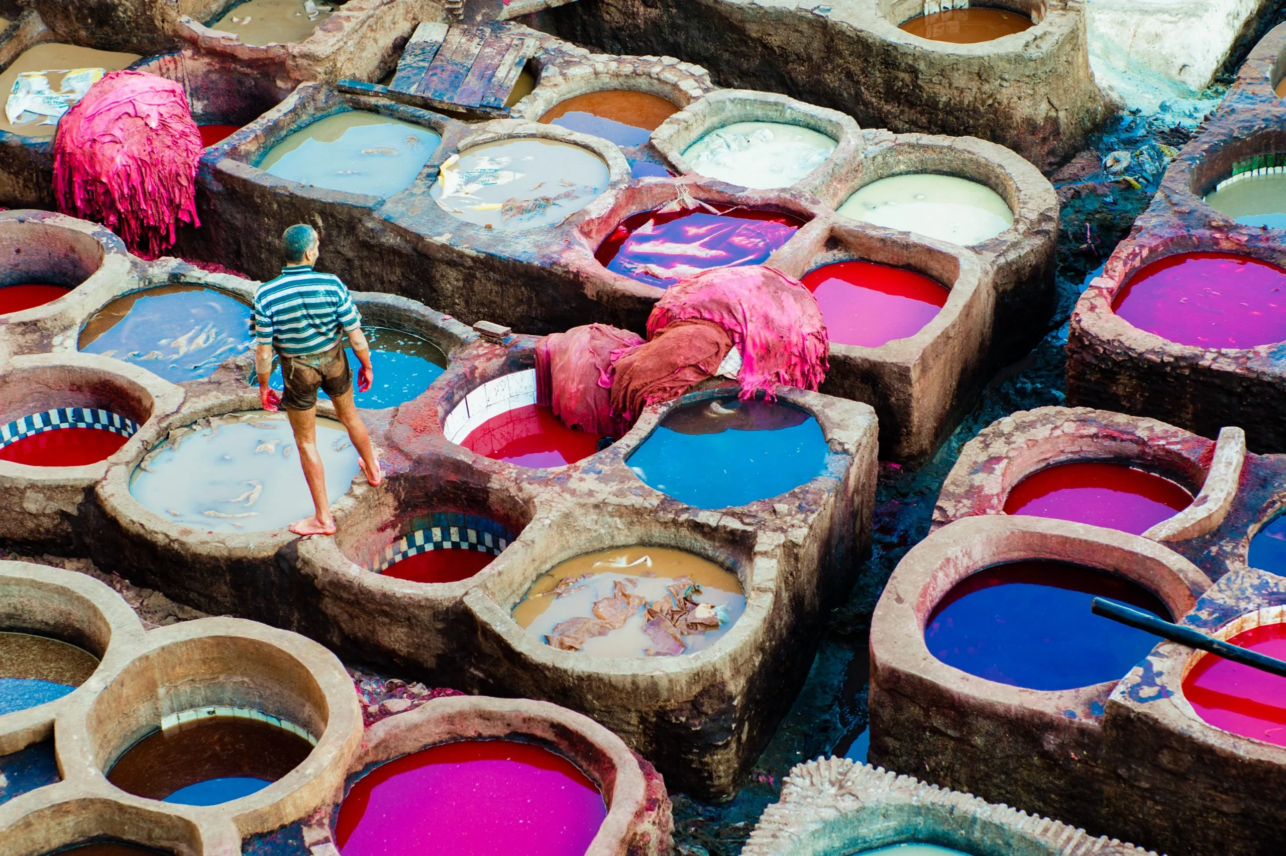 A worker in a striped shirt walks among the dye vats of Fez's Chouara tannery, with bright pink skins draped at the edges.