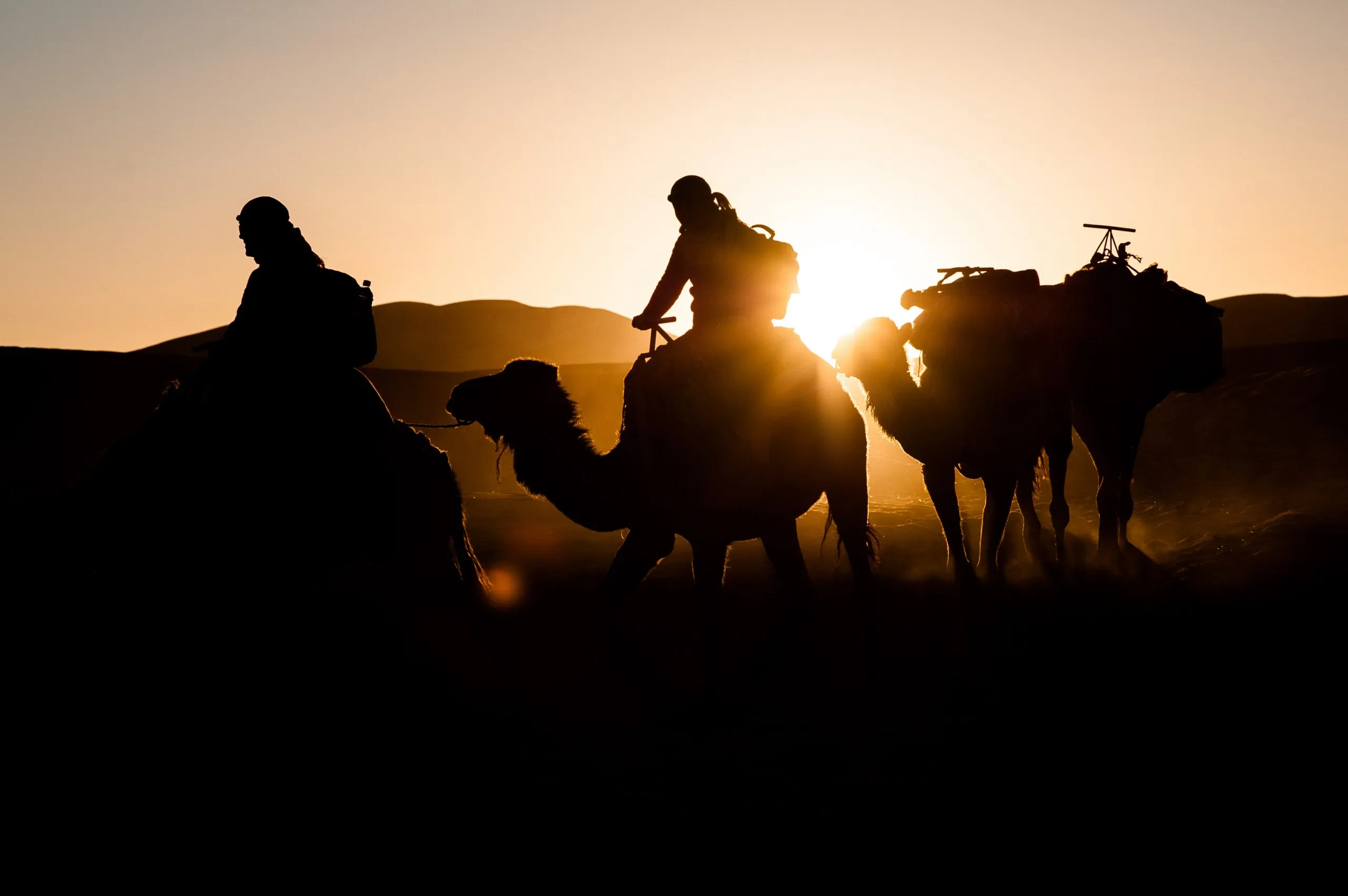 Two camel riders with loaded camels are silhouetted against the Sahara at sunset in Morocco's Erg Chebbi dunes.