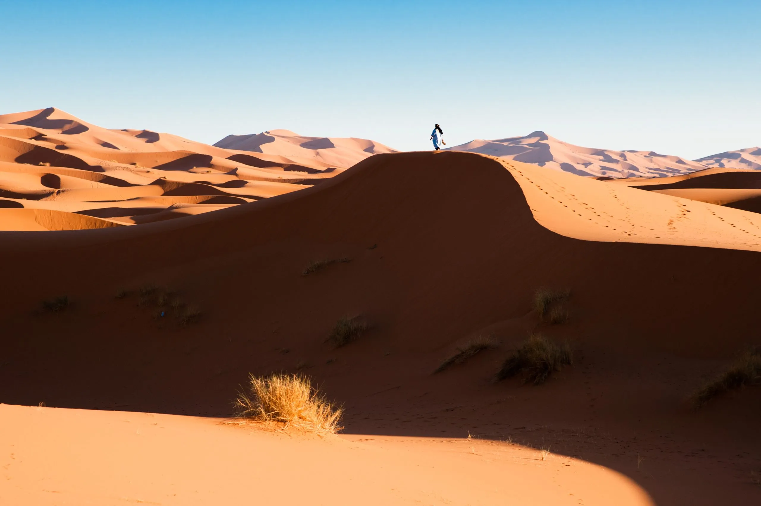 A lone person in blue robes walks the crest of a sand dune deep in Morocco's Erg Chebbi sand sea.