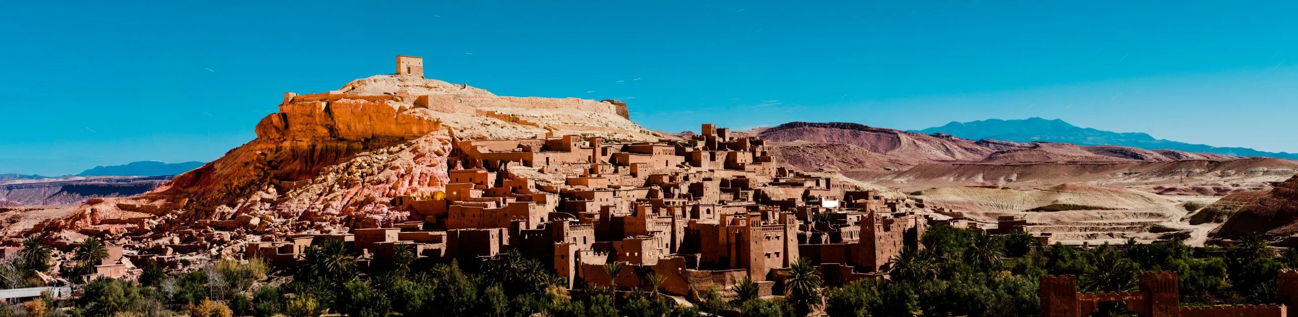 Panoramic view of the red mud-brick fortified village of Aït Ben Haddou in southern Morocco, set against a clear blue desert sky.