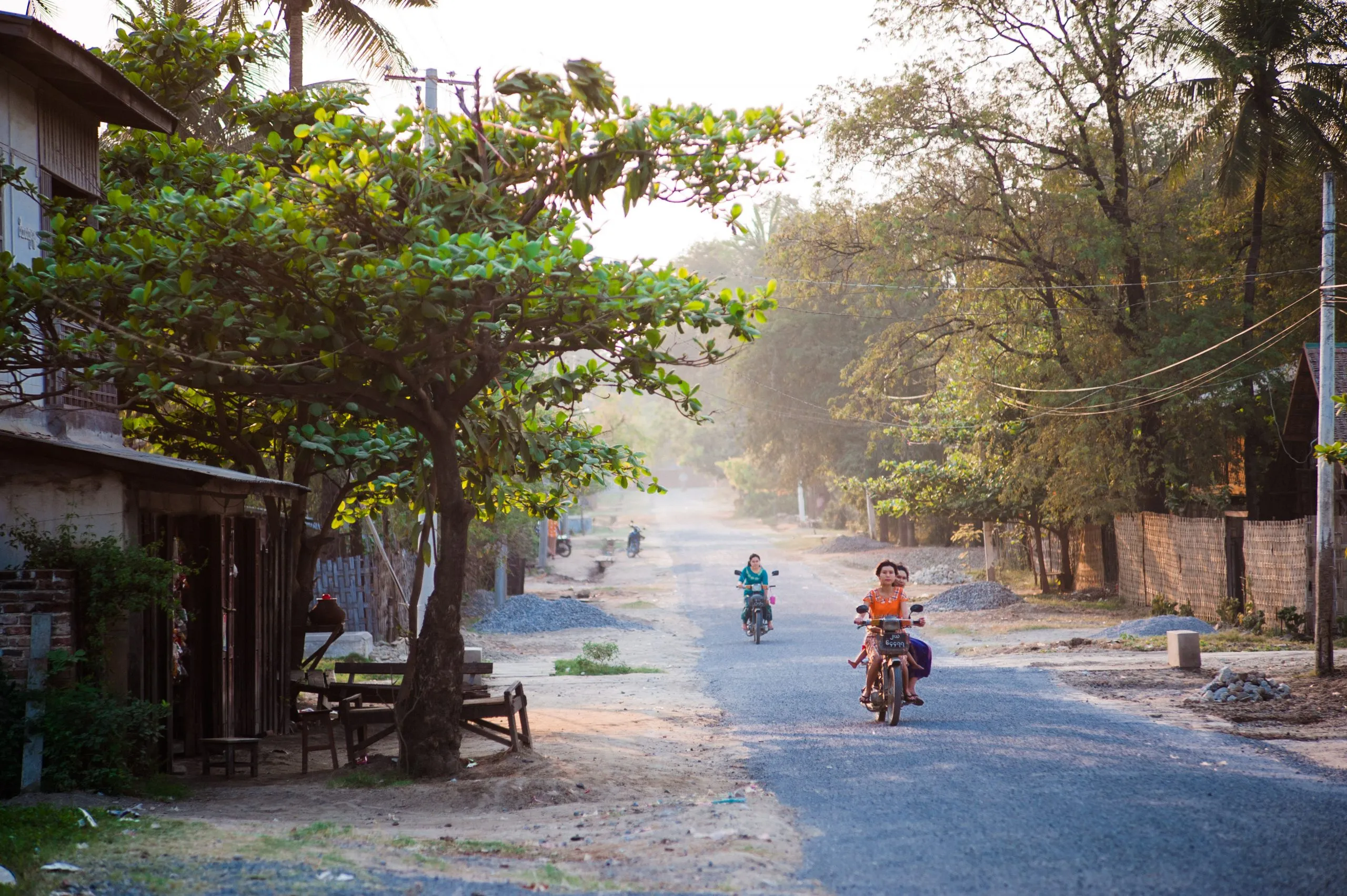 A quiet street at golden hour in Burma, with two motorbikes approaching past a single shade tree.