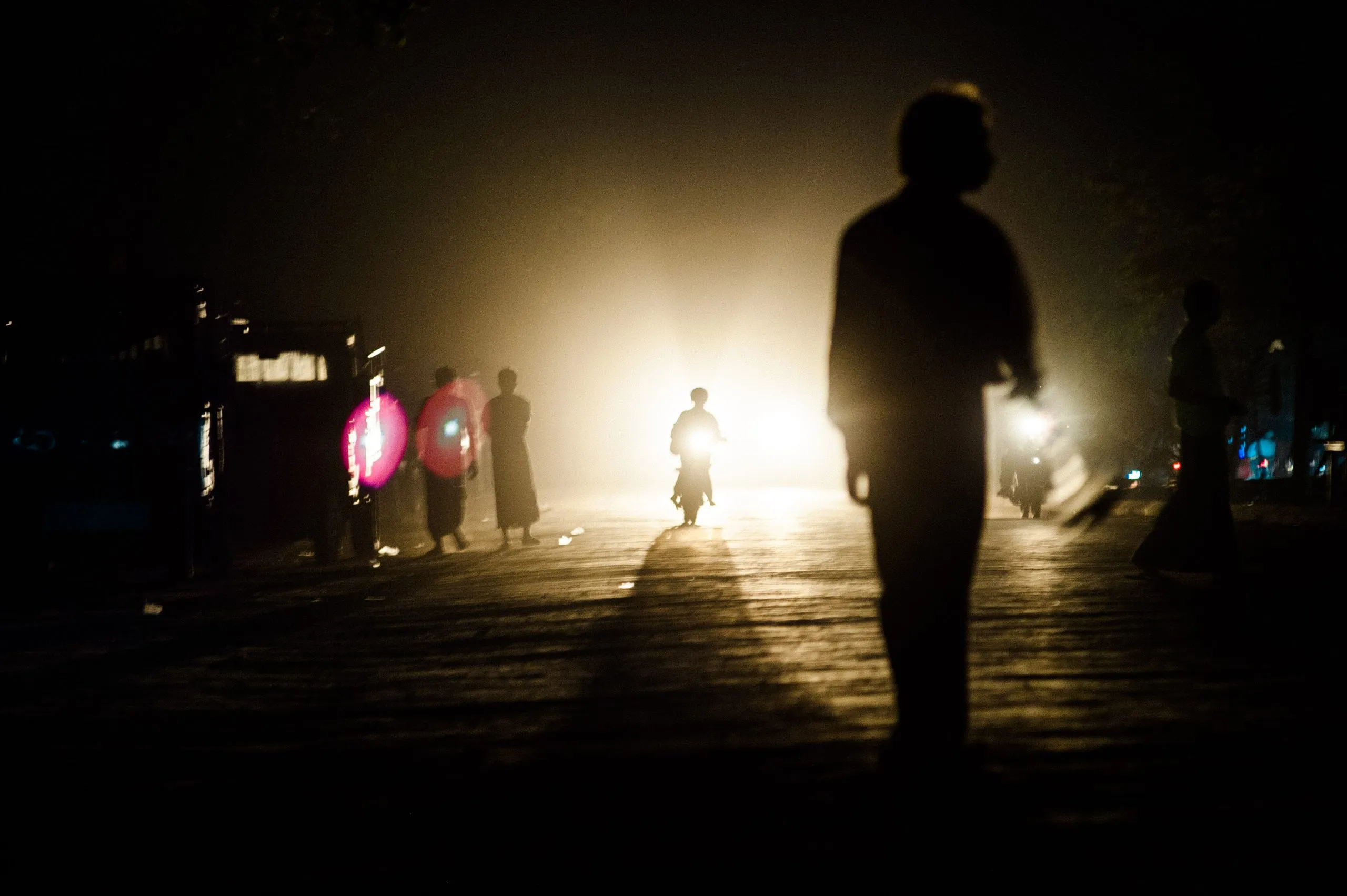 Silhouettes on a foggy Burmese street at night, lit by the headlight of a passing motorbike.