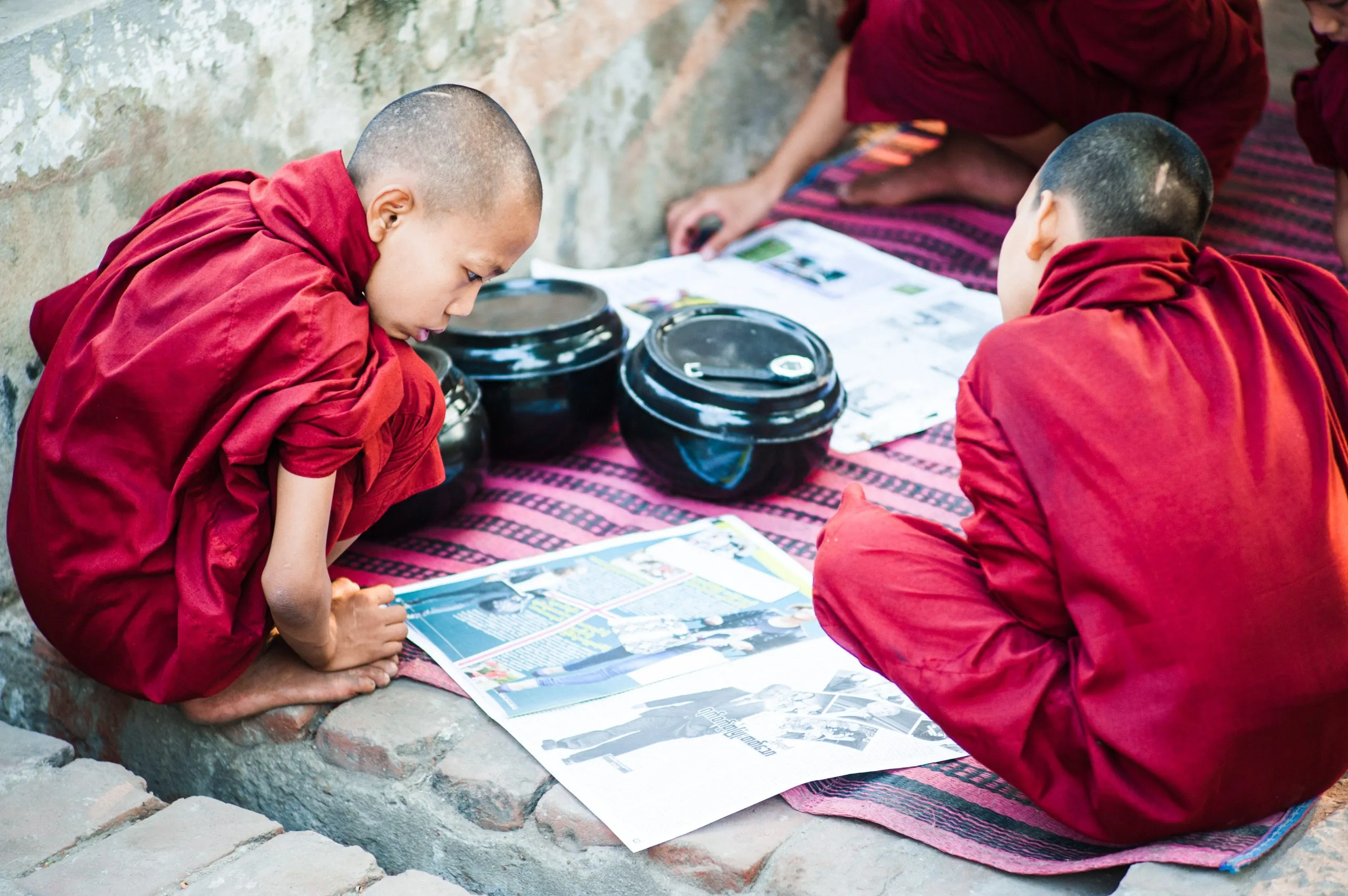 Two young monks read a newspaper on stone temple steps in Bagan, Burma, food bowls set beside them.