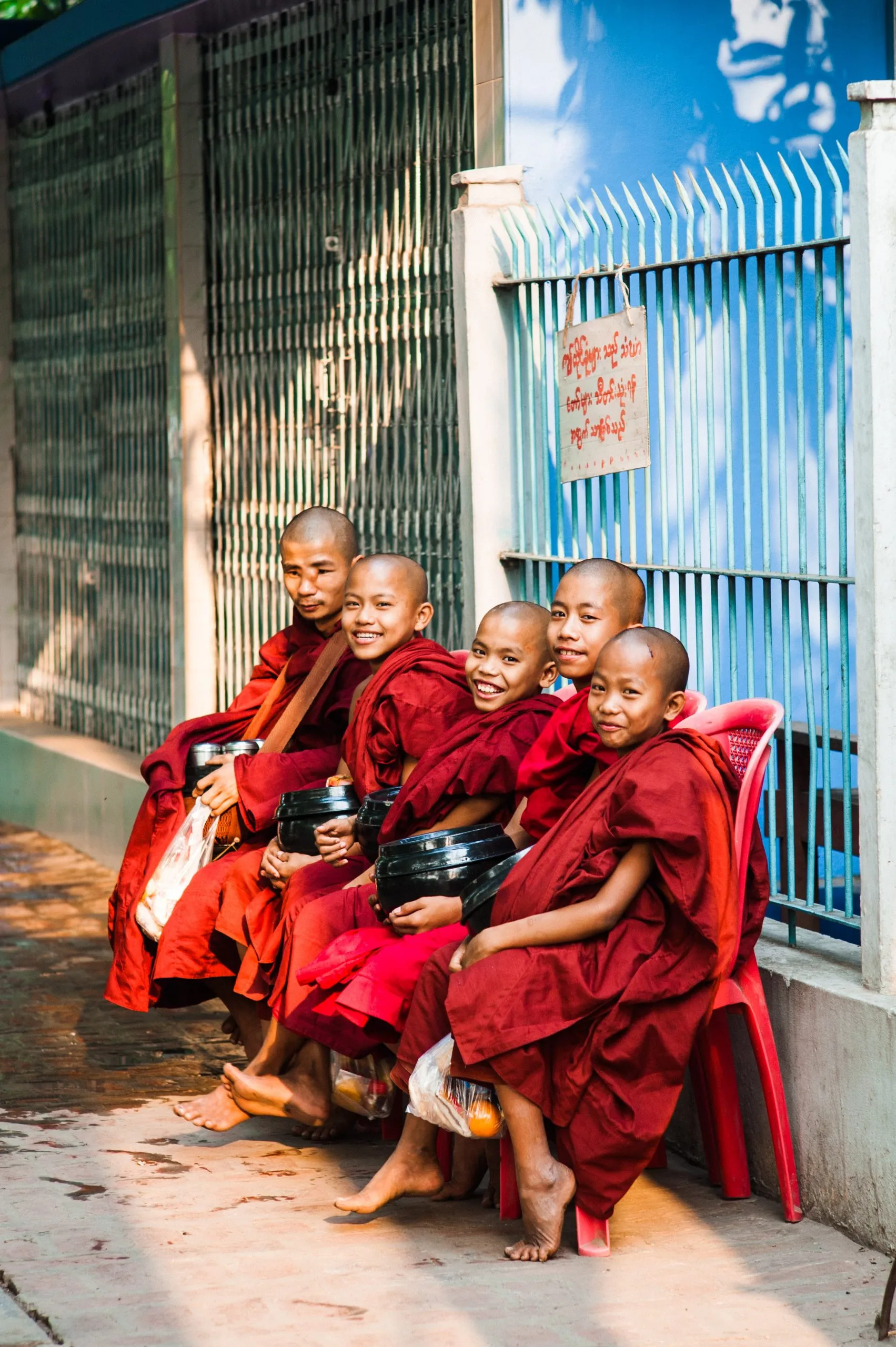 Five young monks crowd together on a small red plastic chair, smiling, on a street in Burma.