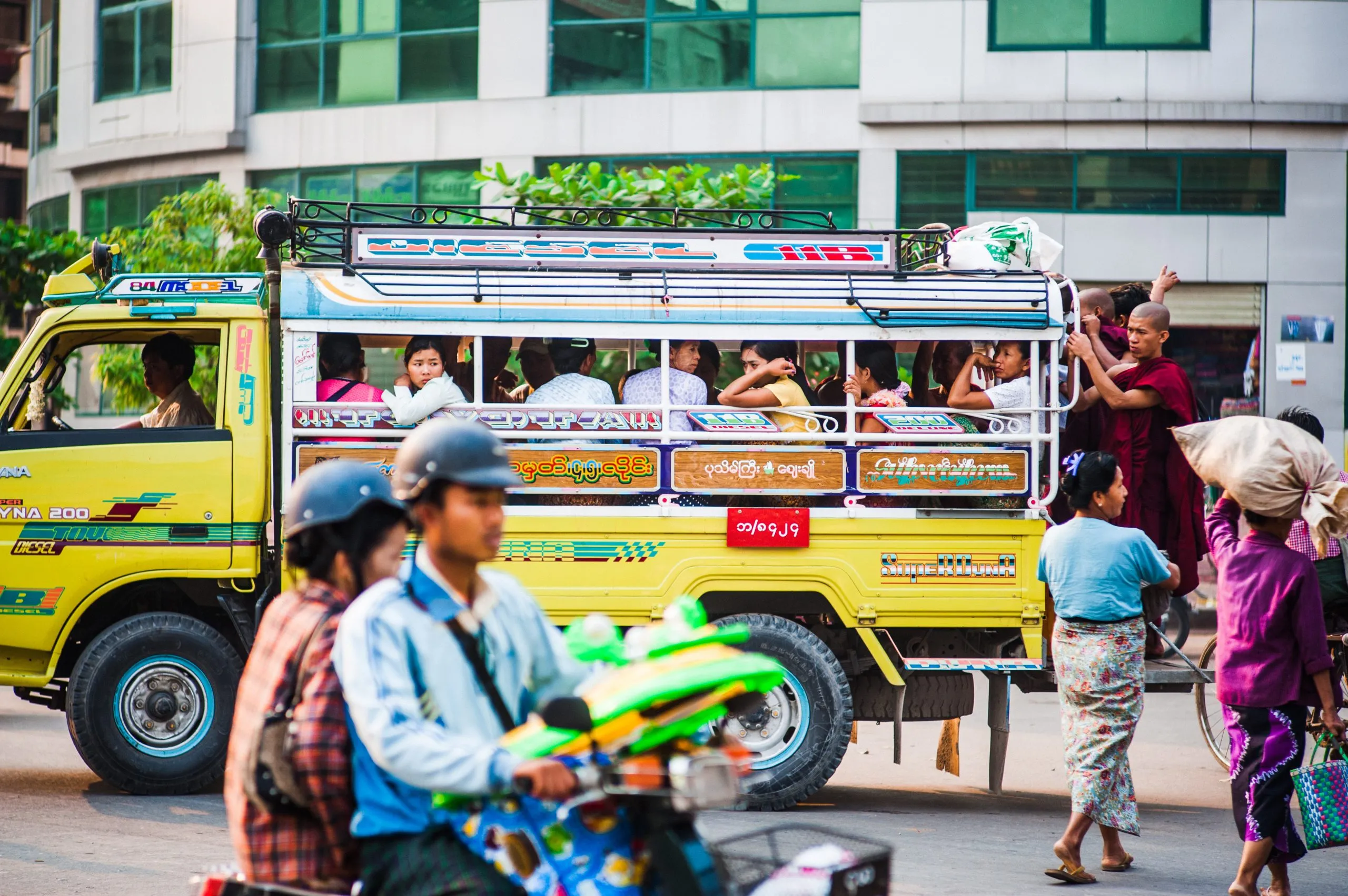 A bright yellow Burmese city bus, packed with passengers, with a motorbike rider passing in front.