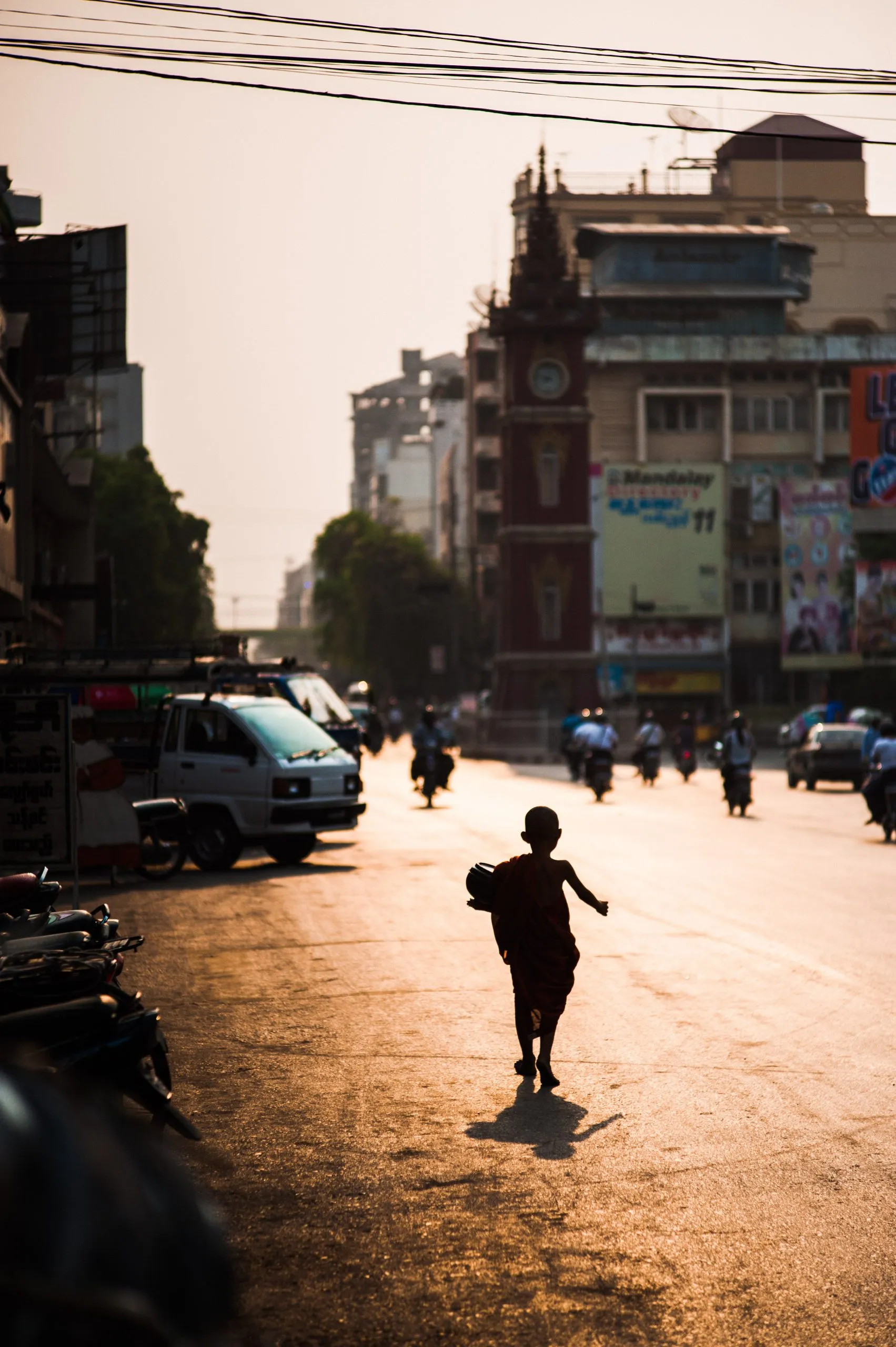 A small monk walks down the middle of a sunset street in Mandalay, the city's clock tower behind in the haze.