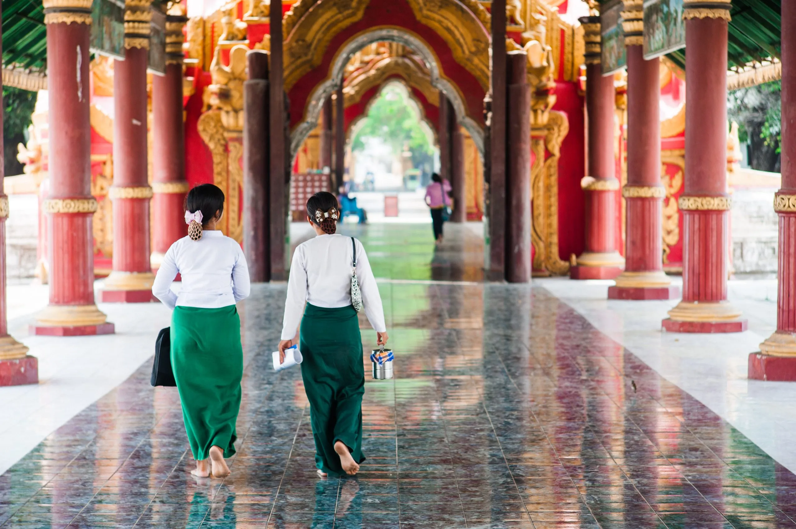 Two young women walk through the red and gold pillared corridor of a Burmese monastery.